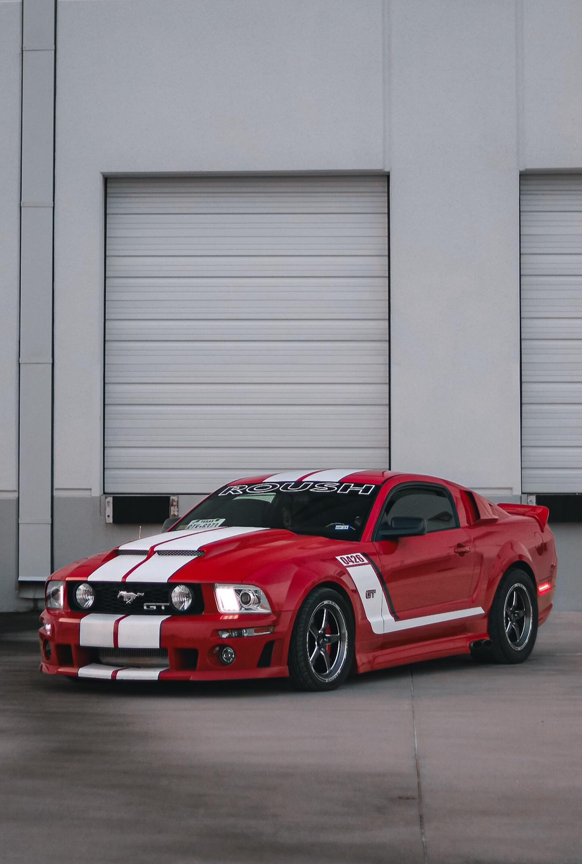 Red Ford Mustang GT with white racing stripes parked in front of a building with large closed white garage doors.