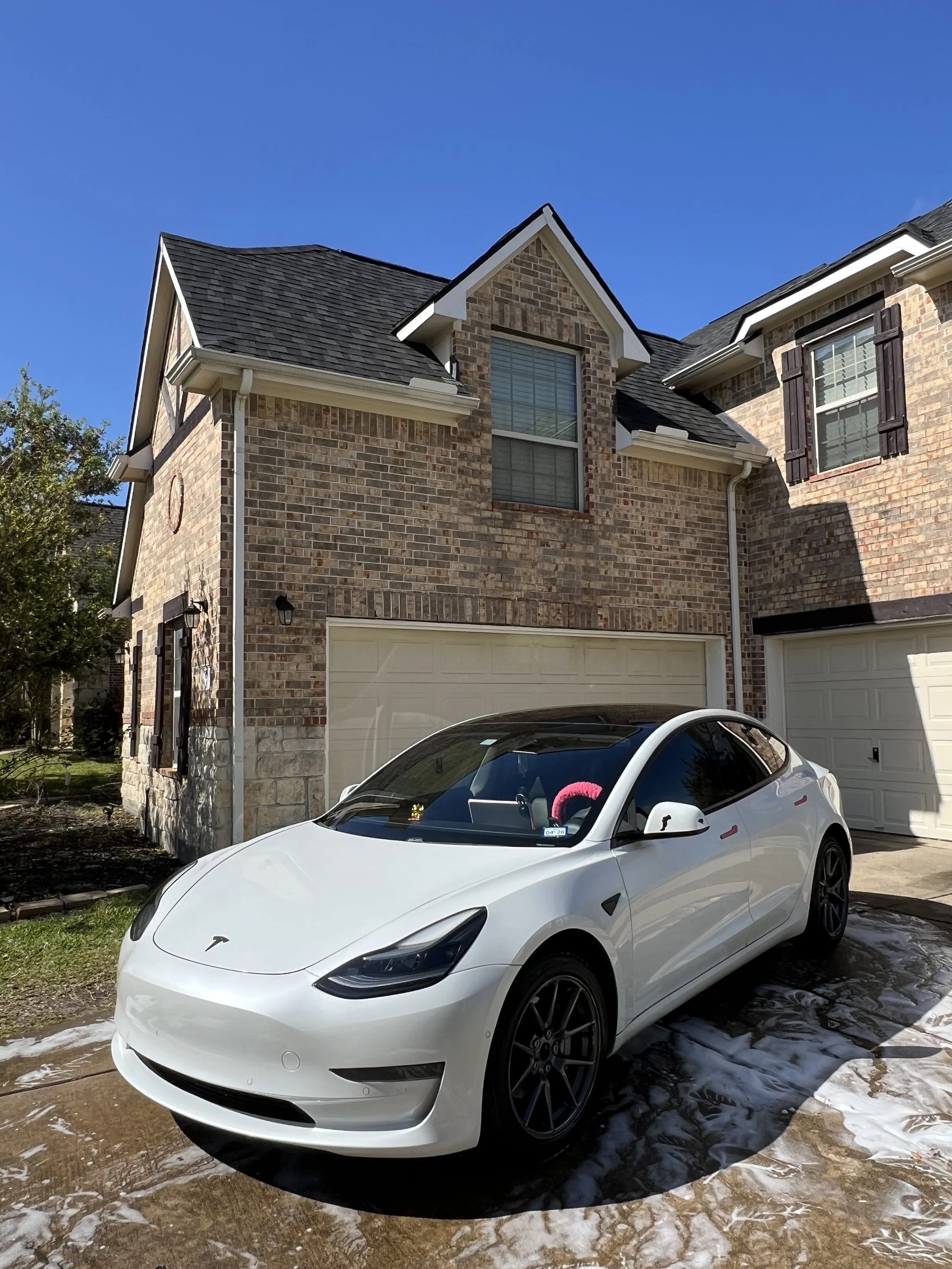A white Tesla Model 3 parked outside a brick house with a two-car garage, on a driveway with soap suds and water from washing the car, under a clear blue sky.