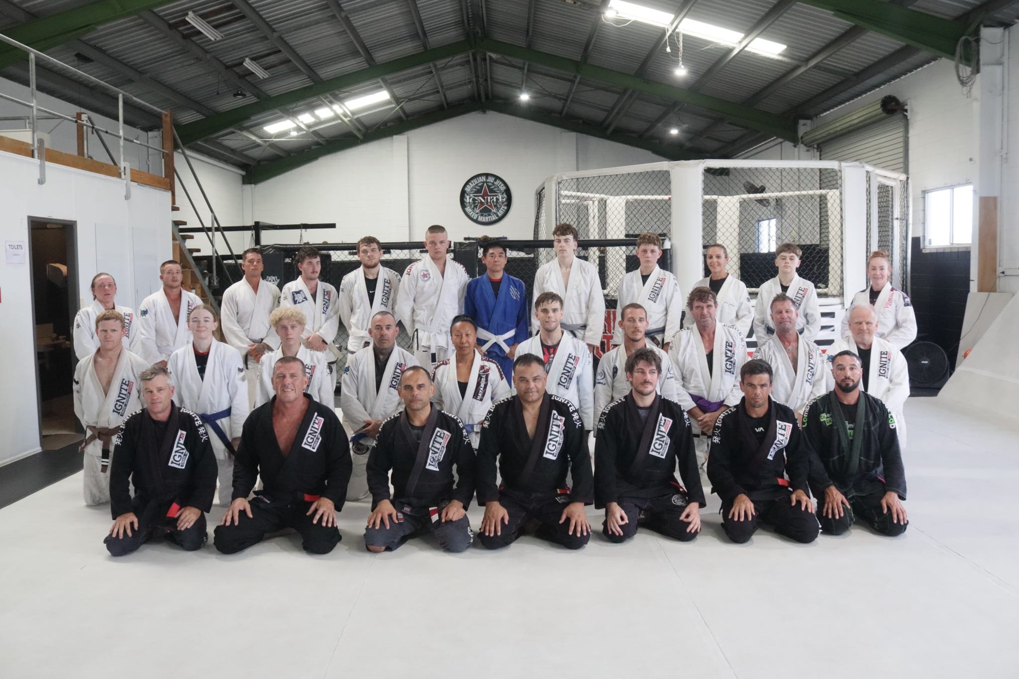 Group photo of martial artists in gi uniforms inside a martial arts training facility with a cage and a sign for Brazilian Jiu Jitsu.