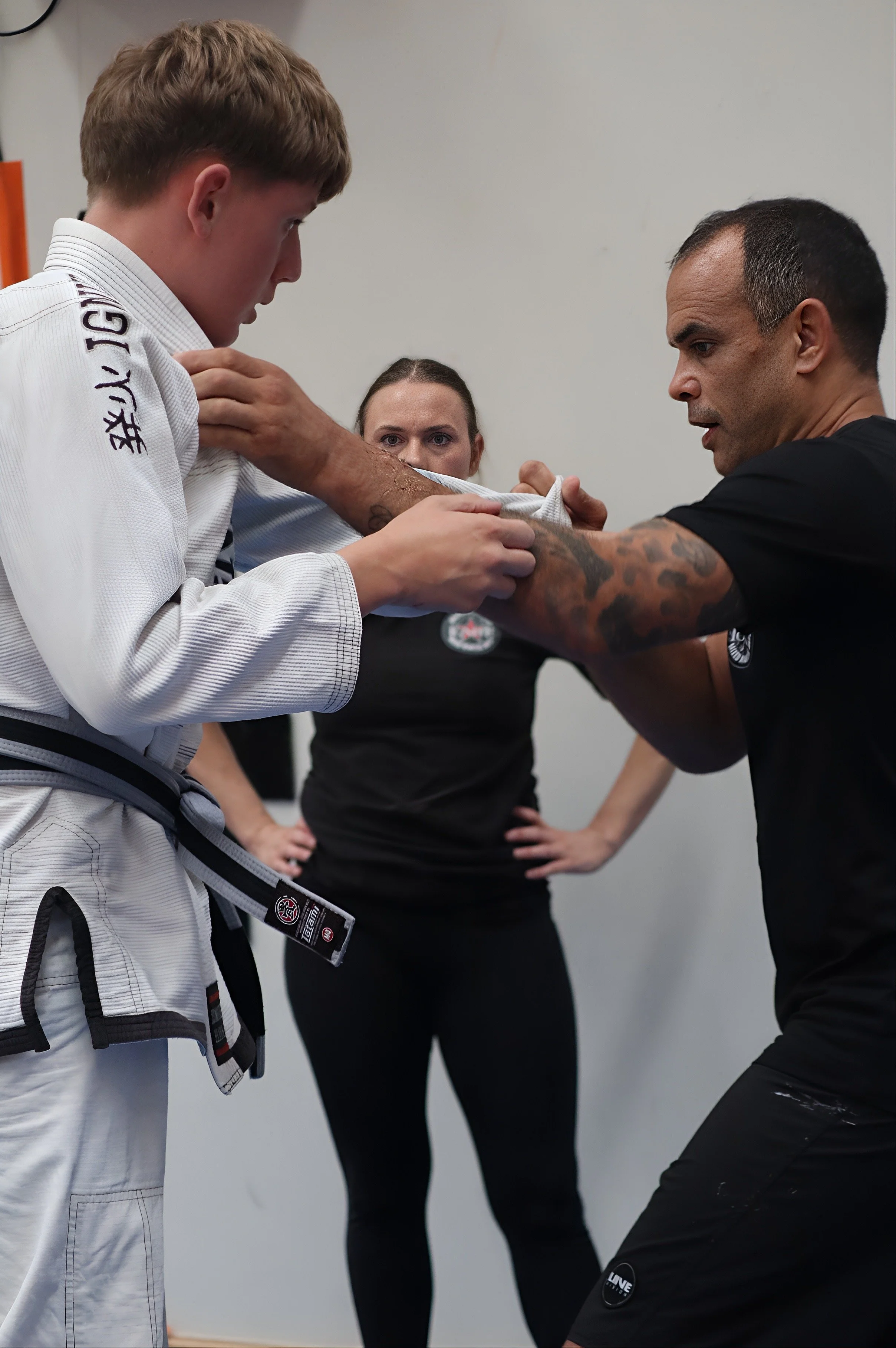 Two men, one in a white martial arts uniform and the other in black athletic clothing, are practicing martial arts together in a training facility. A woman watches in the background.