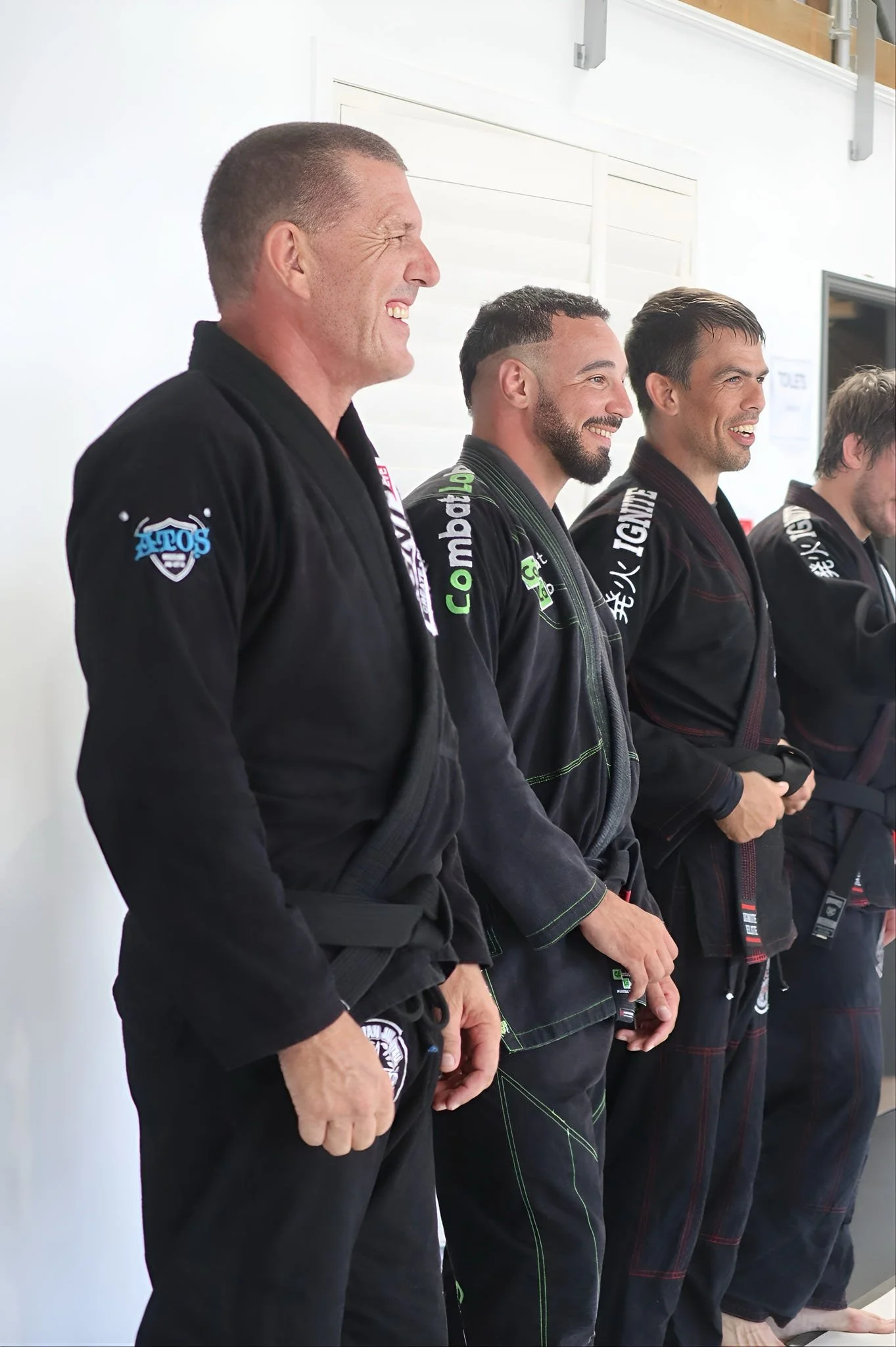 Group of men in martial arts uniforms standing in a row, smiling, against a plain white background.