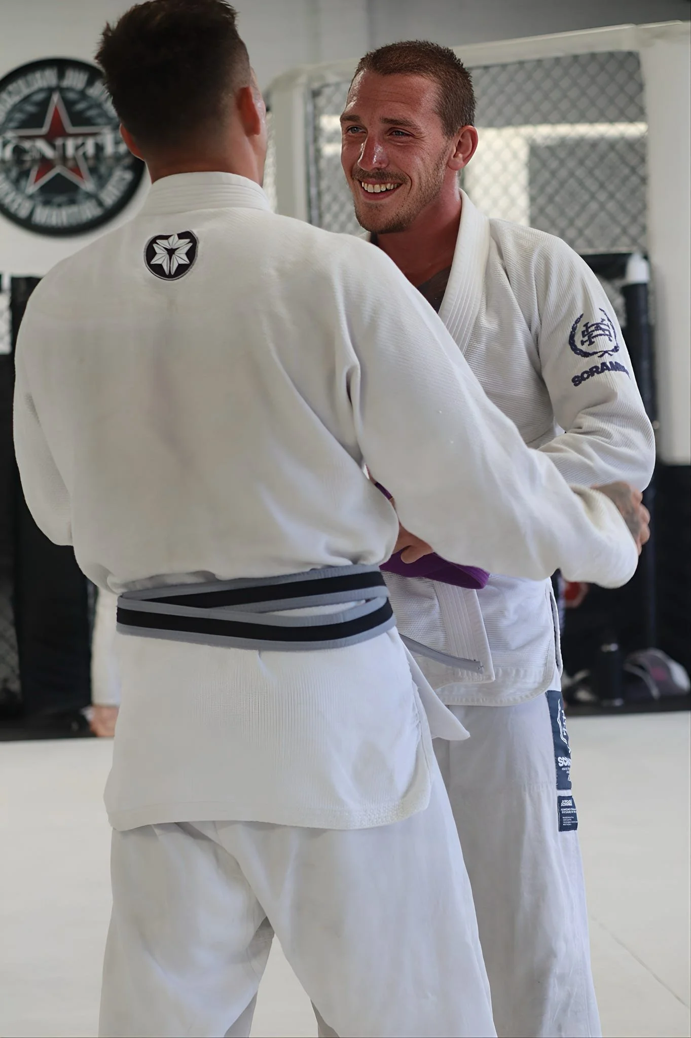 Two men wearing white judo gis with black belts in a martial arts gym, smiling and holding each other.