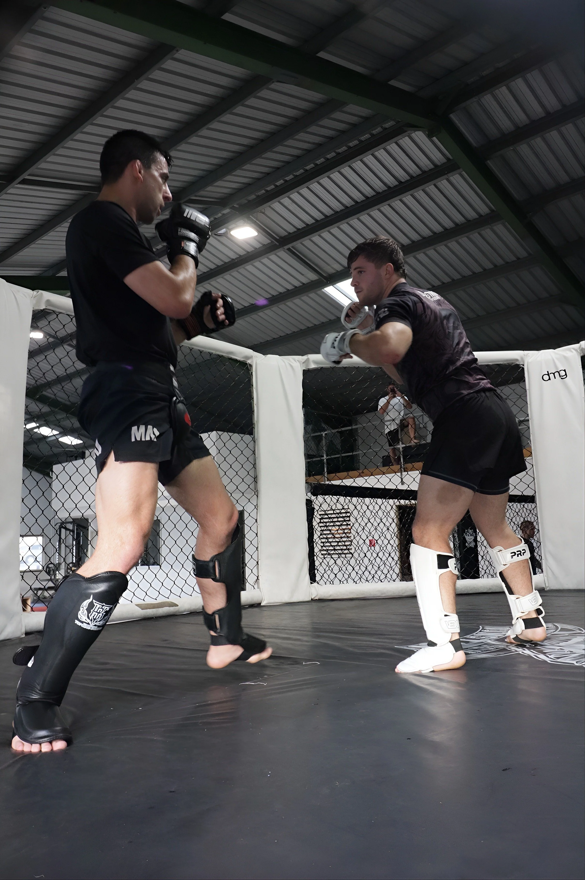 Two male mixed martial arts fighters sparring inside a cage in an indoor gym. One fighter is in black shorts and gloves, the other in black athletic gear and white shin guards. A person is visible in the background taking photos.