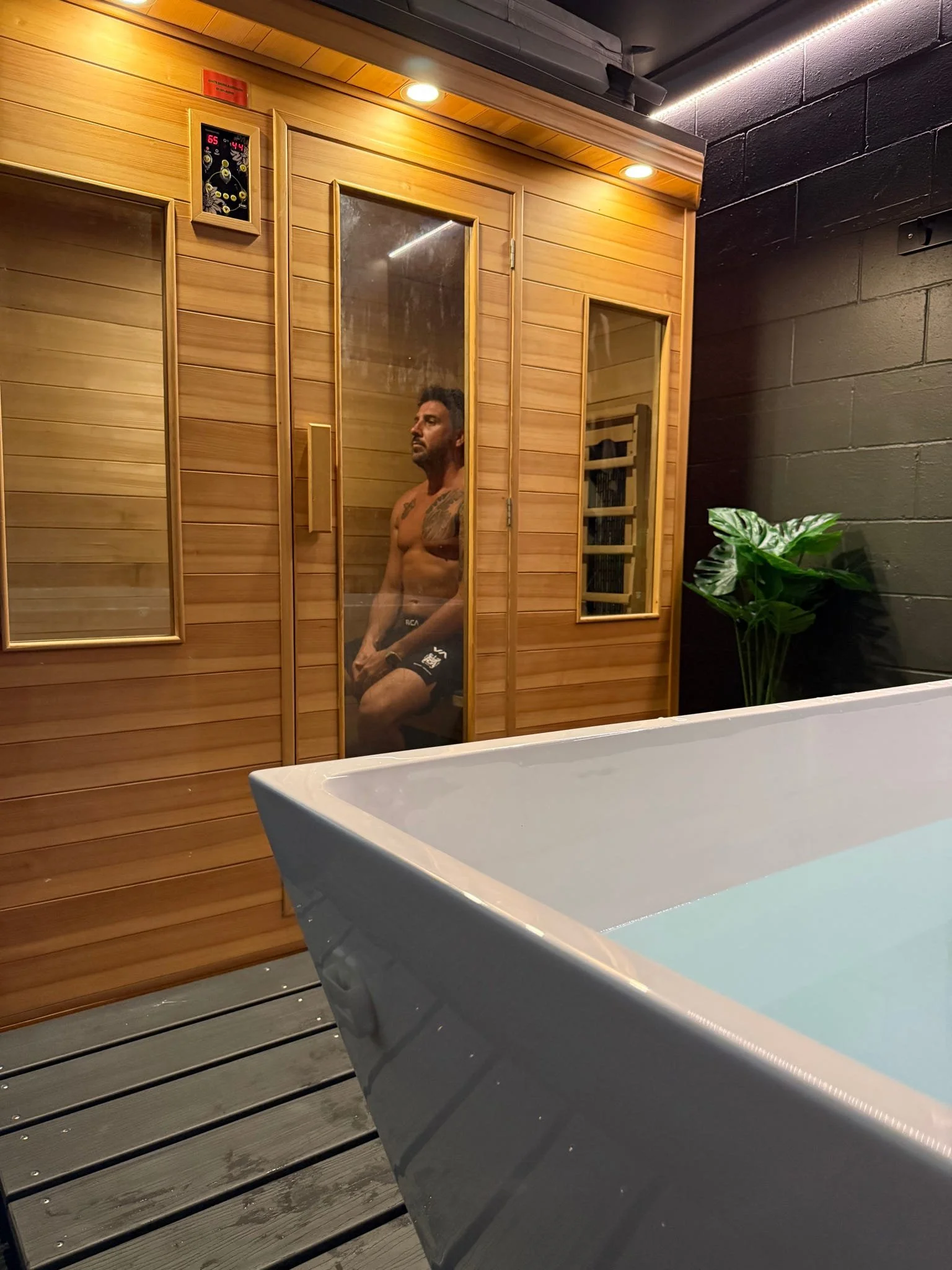 A man sitting in a sauna room behind a glass door. The room is made of wood, and there is a digital temperature control on the wall. Infared sauna available to members and the public through our booking system.