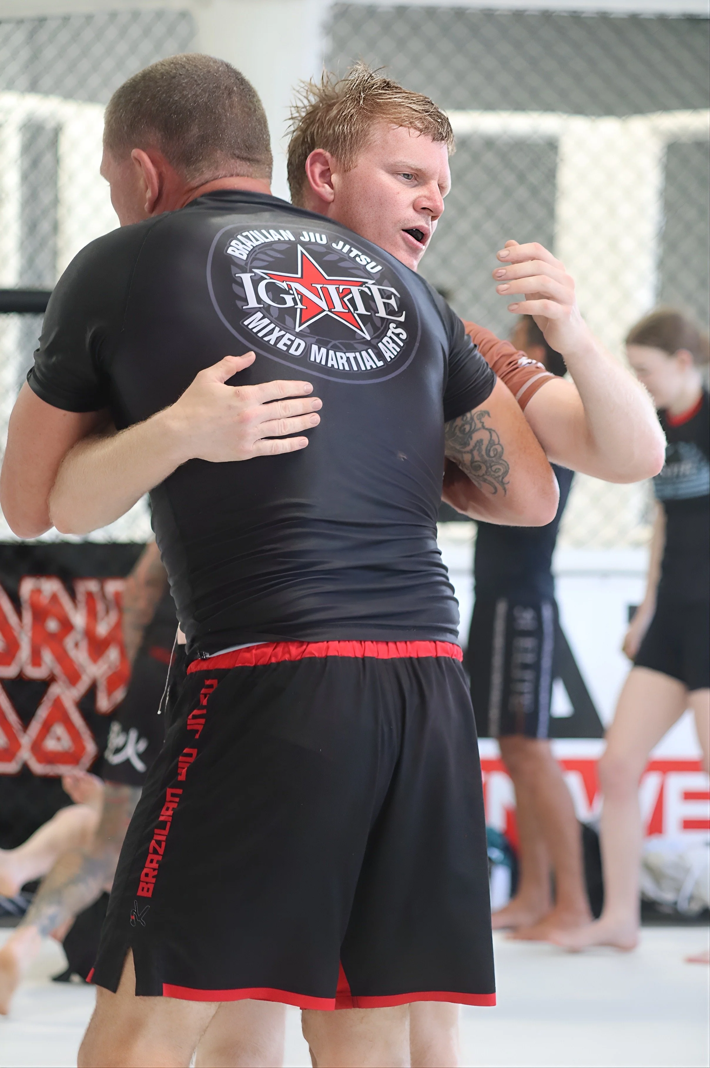 Two men practicing Brazilian Jiu Jitsu in a training gym. One man is hugging the other, who has open-mouth facial expression. They are wearing BJJ uniforms with logos and shorts. Other people are practicing in the background.