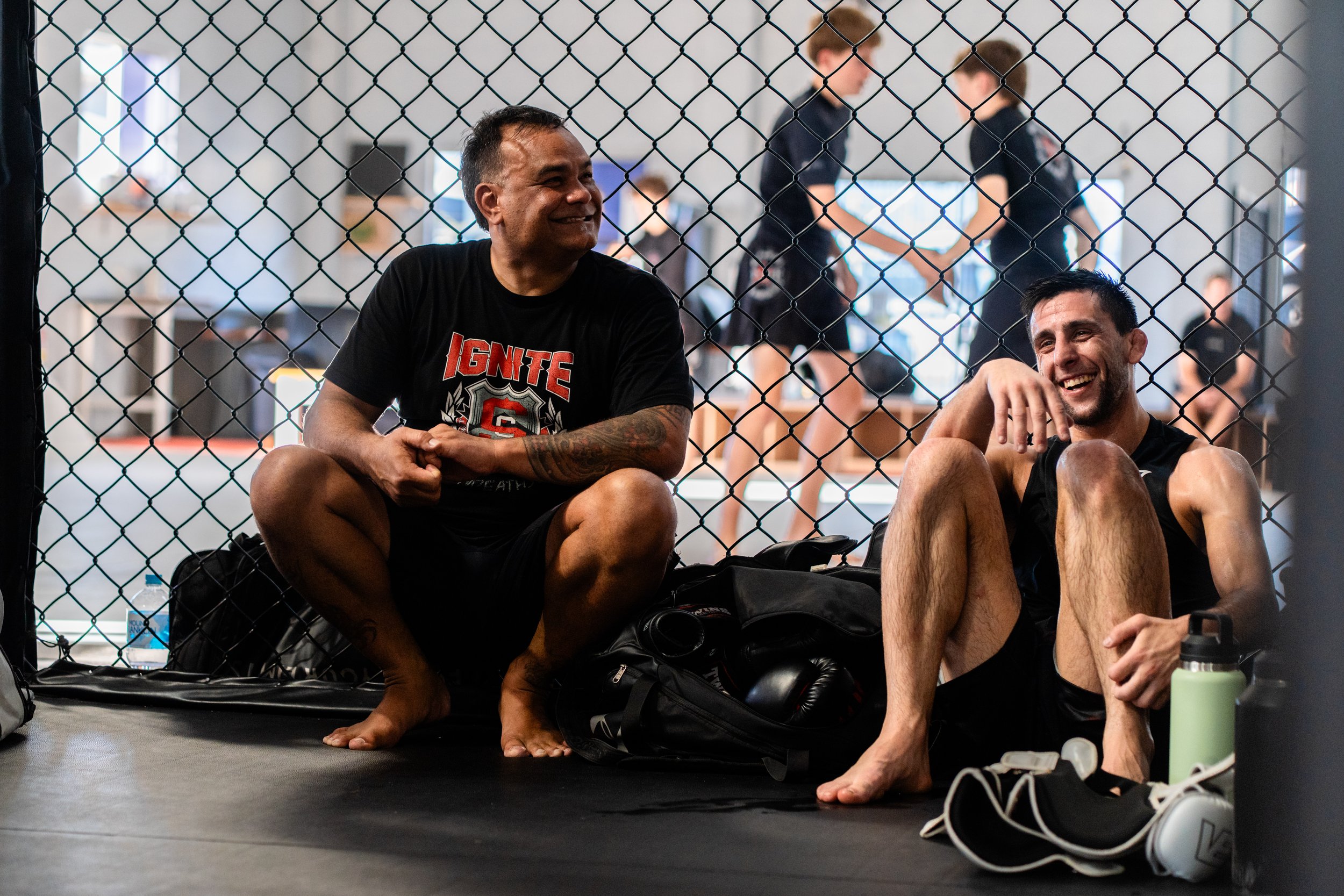 Two men sitting on the floor inside a behind-the-fence fighting cage, smiling and laughing, with three women in the background inside a gym, and various workout equipment around.