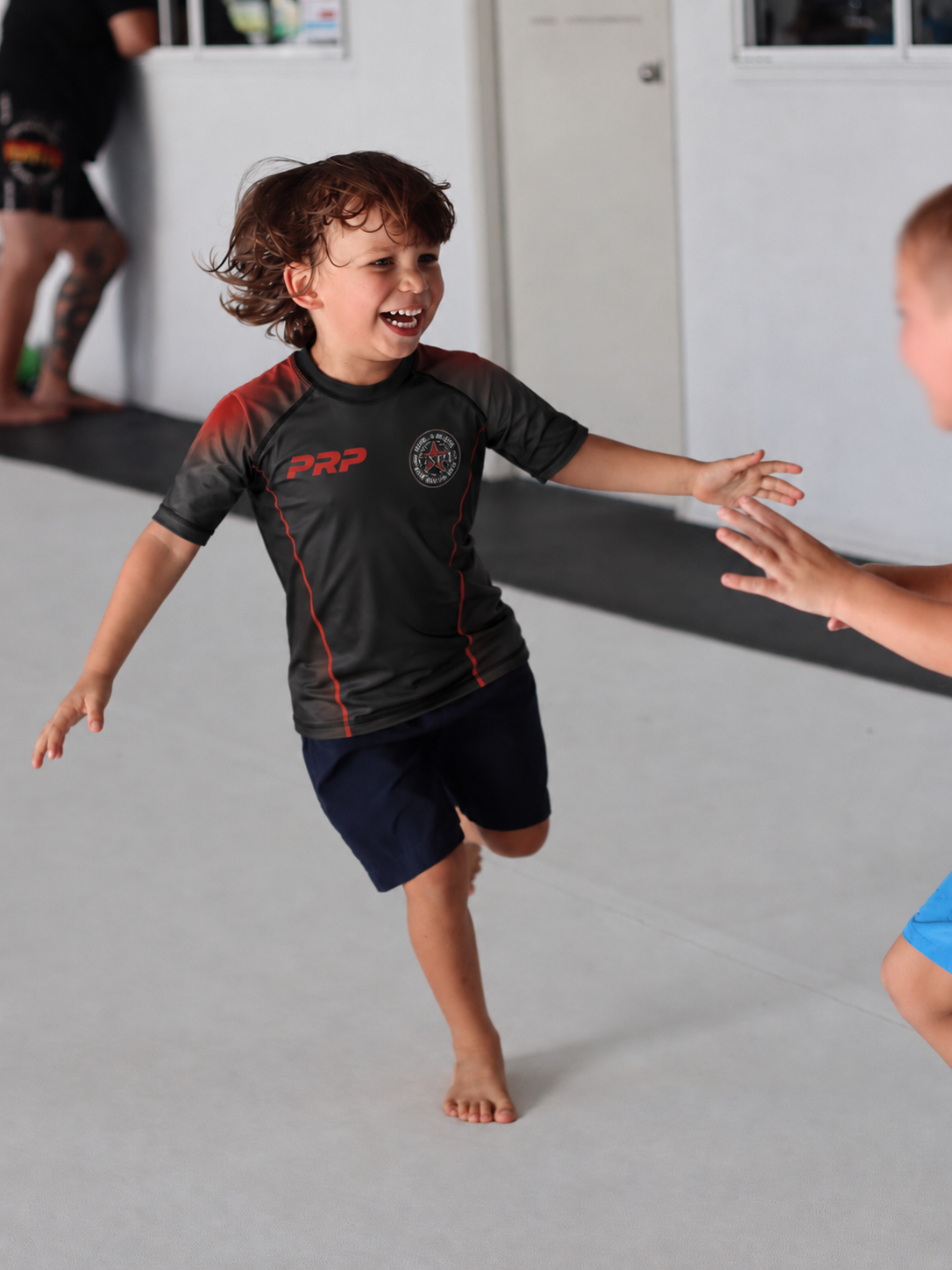 Two young children are practicing Brazilian Jiu-Jitsu, smiling and reaching out to each other, in a martial arts gym.