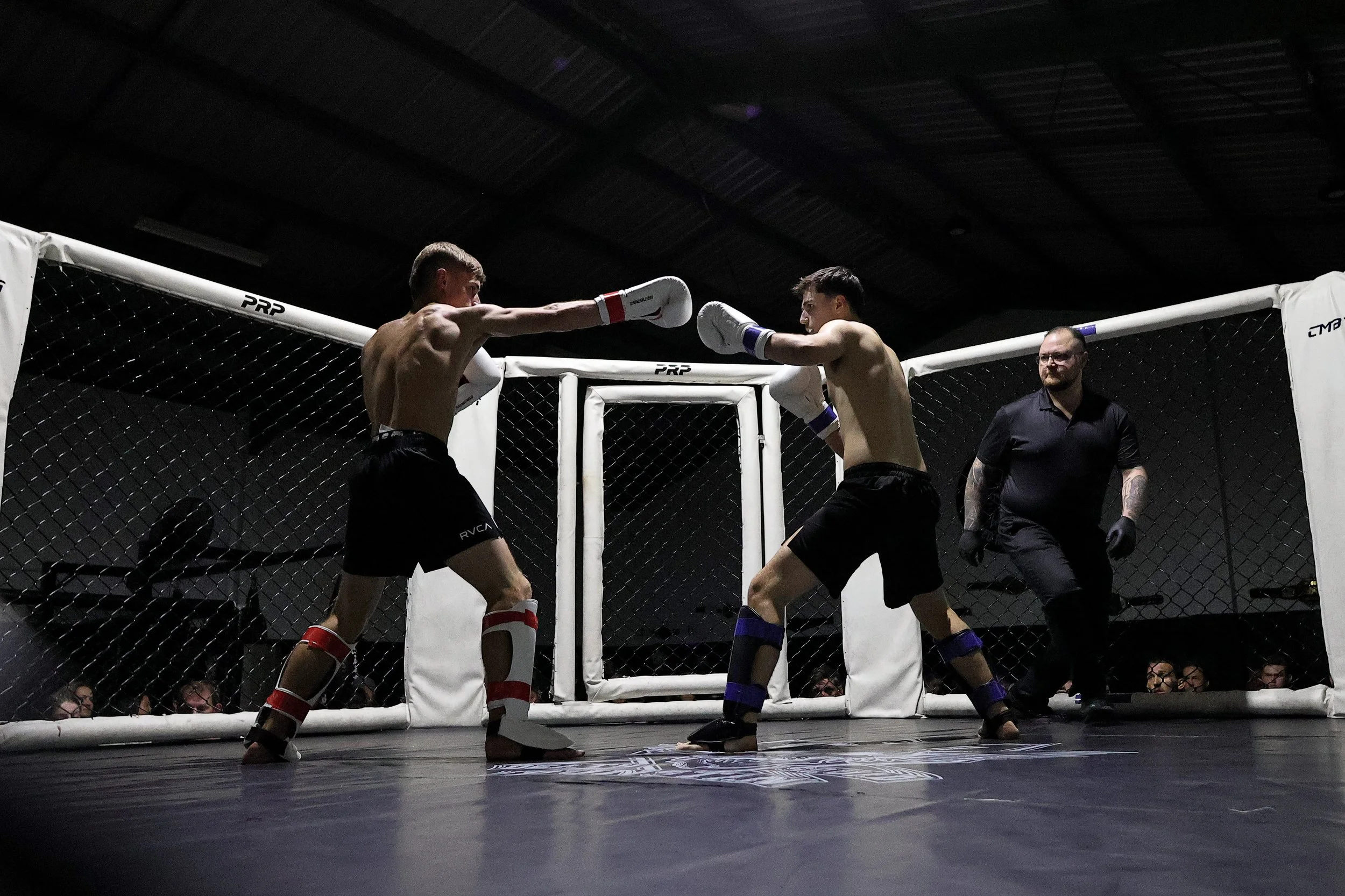 Two male fighters in a mixed martial arts match inside a cage, one throwing a punch at the other's face, with a referee observing nearby, in a dark indoor arena. Great training facility with multiple training spaces and gear.