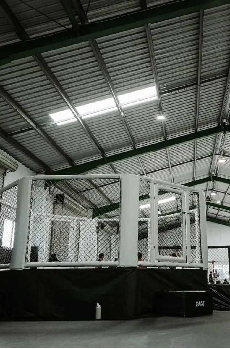 Indoor mixed martial arts fighting cage inside a gym with a metal roof, surrounded by a black padded floor and a few people visible in the background.