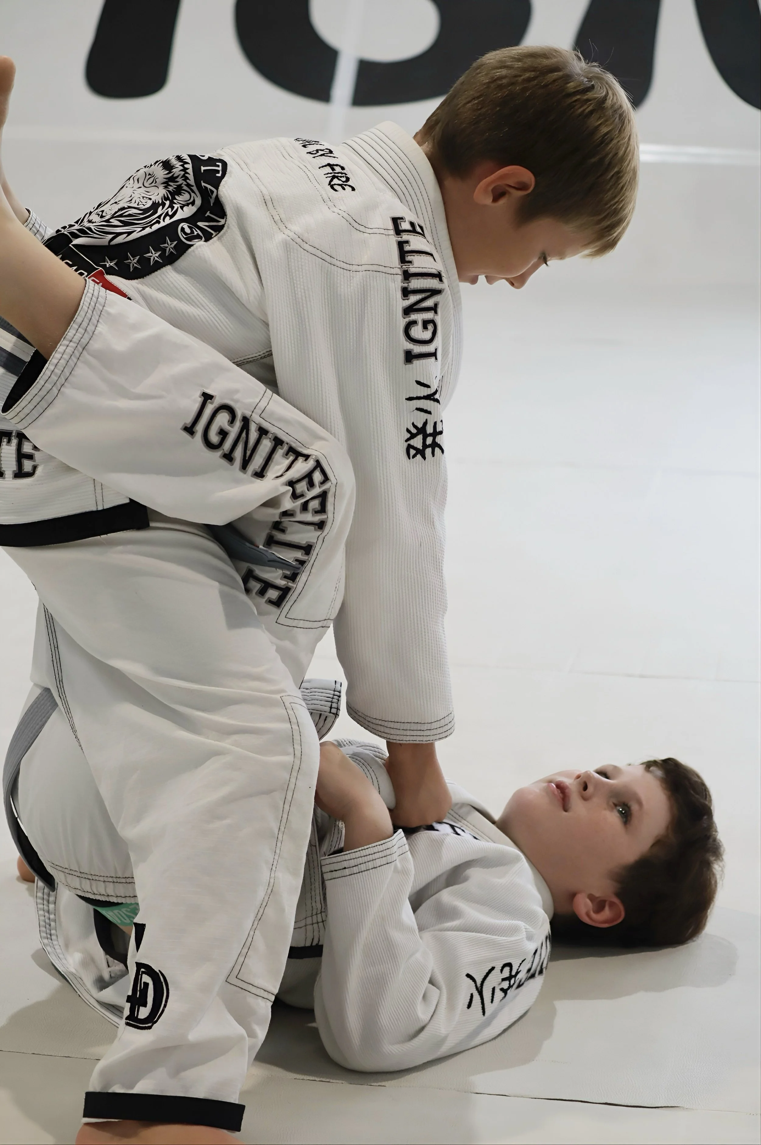 Two young boys in white Brazilian Jiu-Jitsu gis practicing grappling on a white mat. One boy is on his back, looking upward, while the other boy is on top, leaning forward with an intense focus. The scene is in a martial arts training facility.