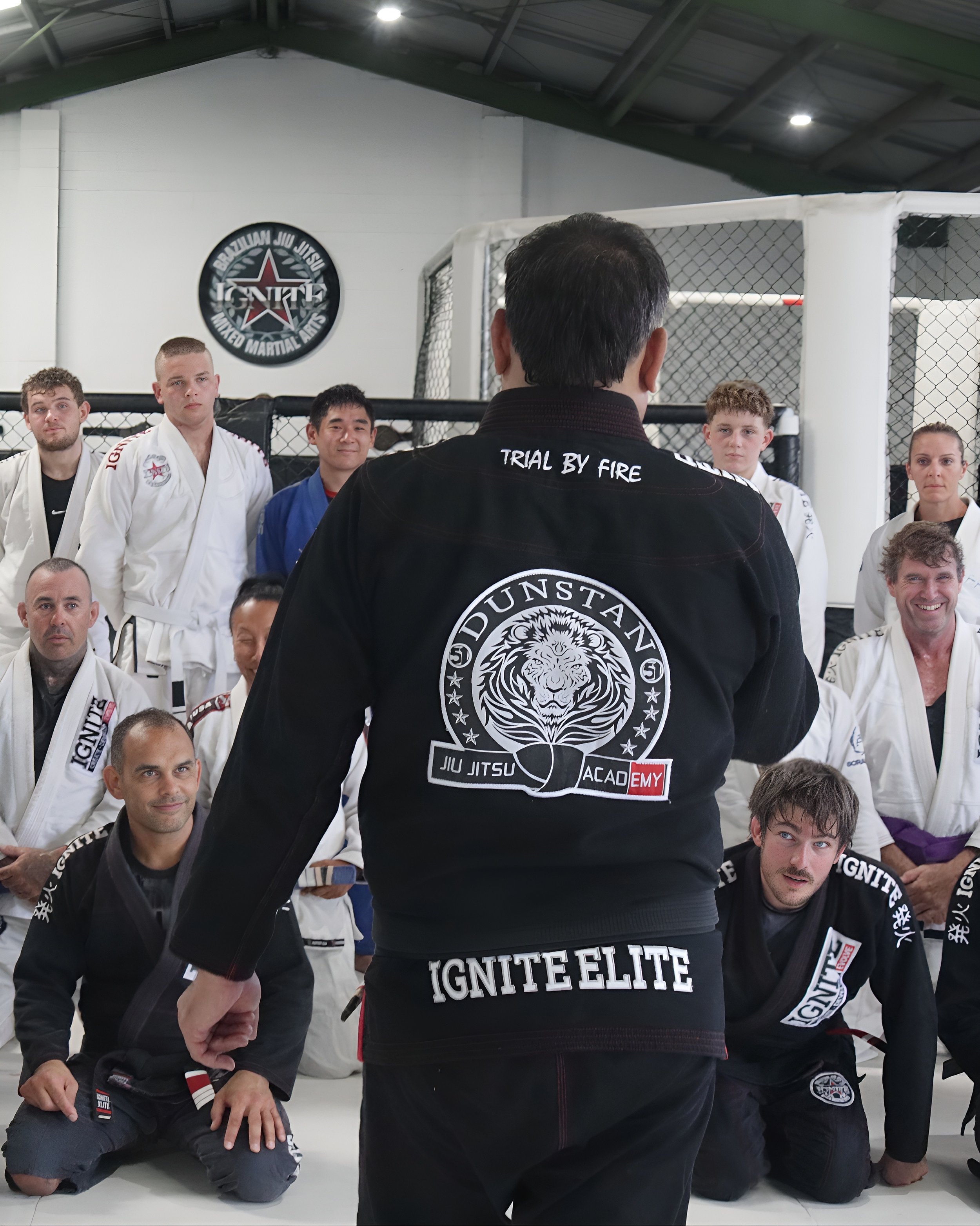 Martial arts class in a gym with students in gis sitting on the mat, listening to instructor who is wearing a black gi with 'Trial by Fire' and 'Ignite Elite' logos, inside a cage MMA training area.