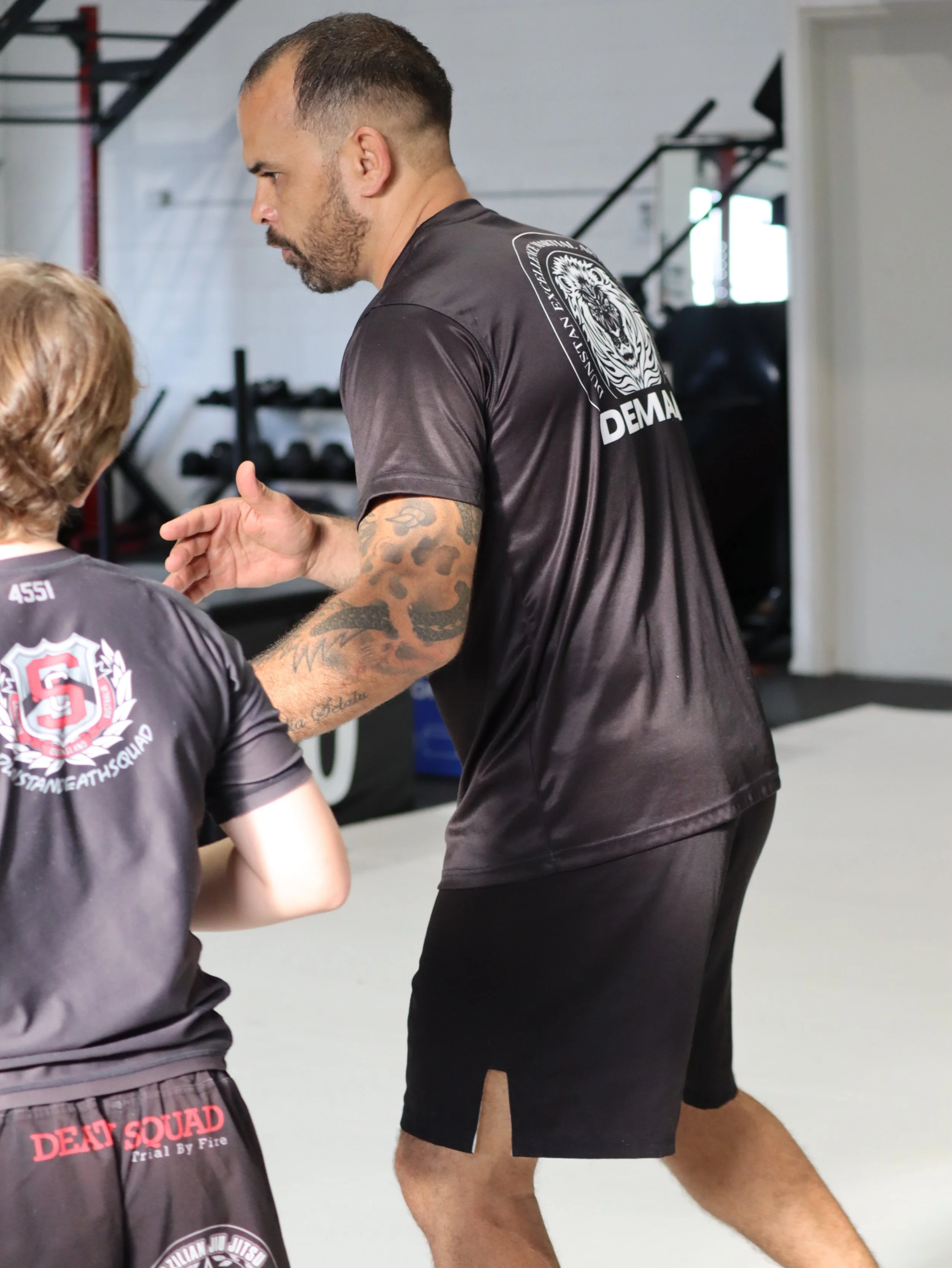 Two men in athletic gear training in a gym. The man in the foreground has tattoos on his arm and is wearing black shorts with a side slit, while the man in the background is wearing a black T-shirt with a lion graphic and the words 'DEMI LAT'.