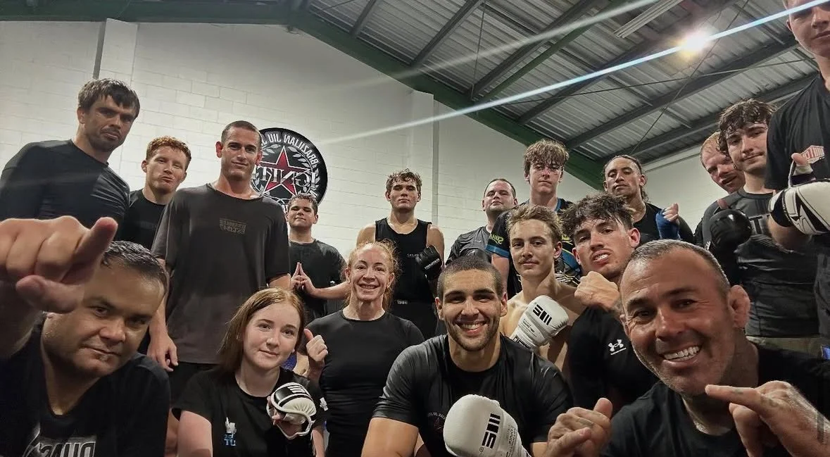 Group of people, including fighters and trainers, in a gym, some wearing boxing gloves, posing for a photo after a workout or fight. They are smiling and making fist gestures.