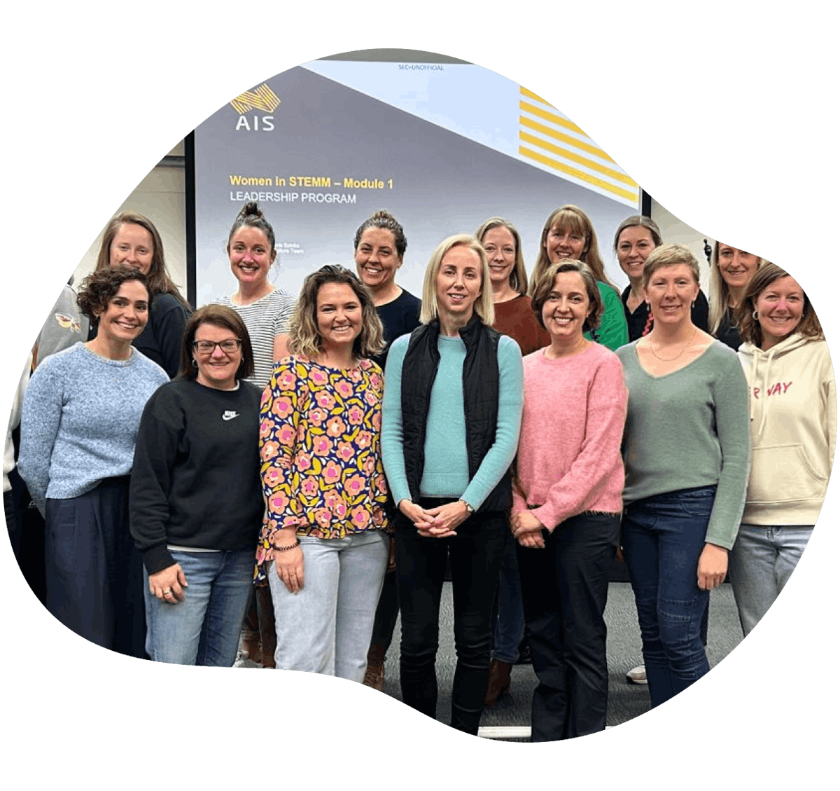 A group of women stand infront of a projector screen, posing for a group photo to celebrate the Women in STEMM Leadership Develop Program delivered by Nobis.