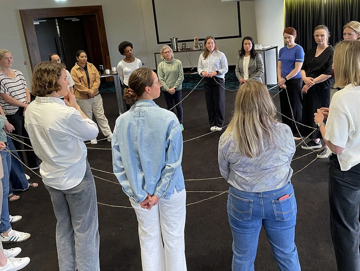 A group of women stand in a circle connected by criss-crossed strings during a team development activity.