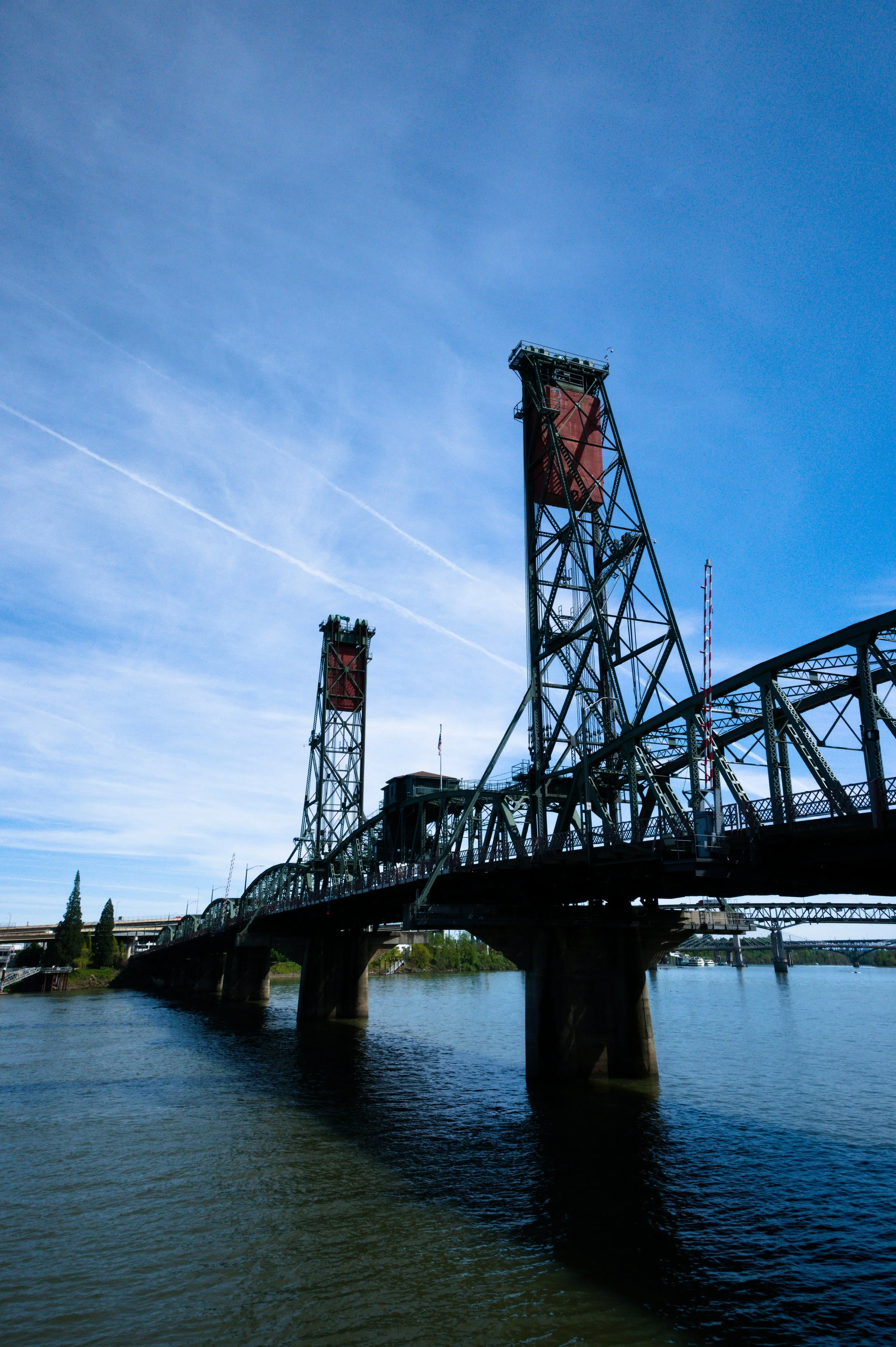 An industrial bridge with metal towers spanning over a body of water in a cityscape, under a blue sky with light clouds and contrails.