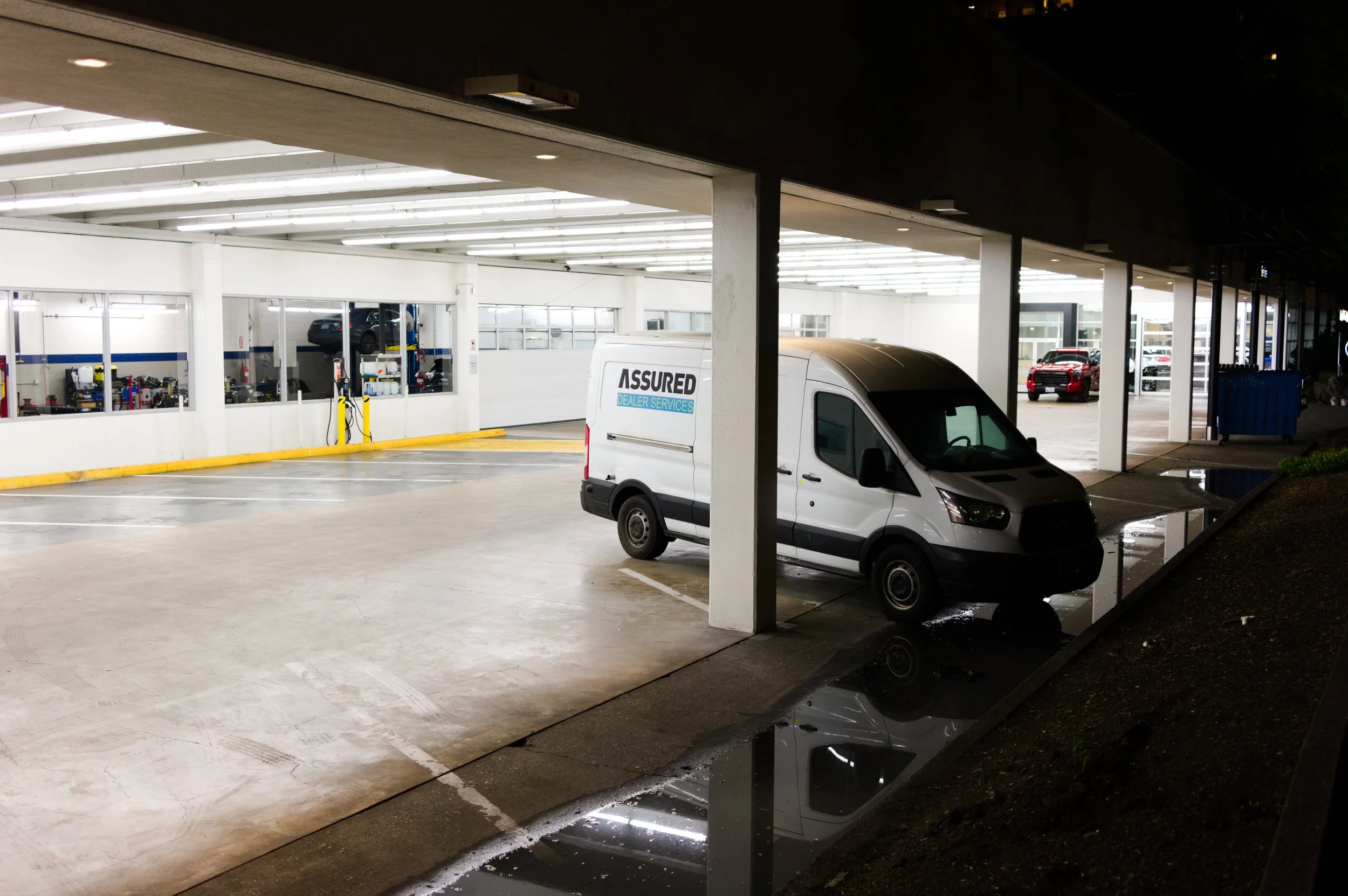 A car repair shop with a white van parked outside, with the shop front showing a sign that says 'Assured Dealer Services.' Inside, there are cars being serviced, and tools and equipment are visible through the windows. The scene appears to be taken a