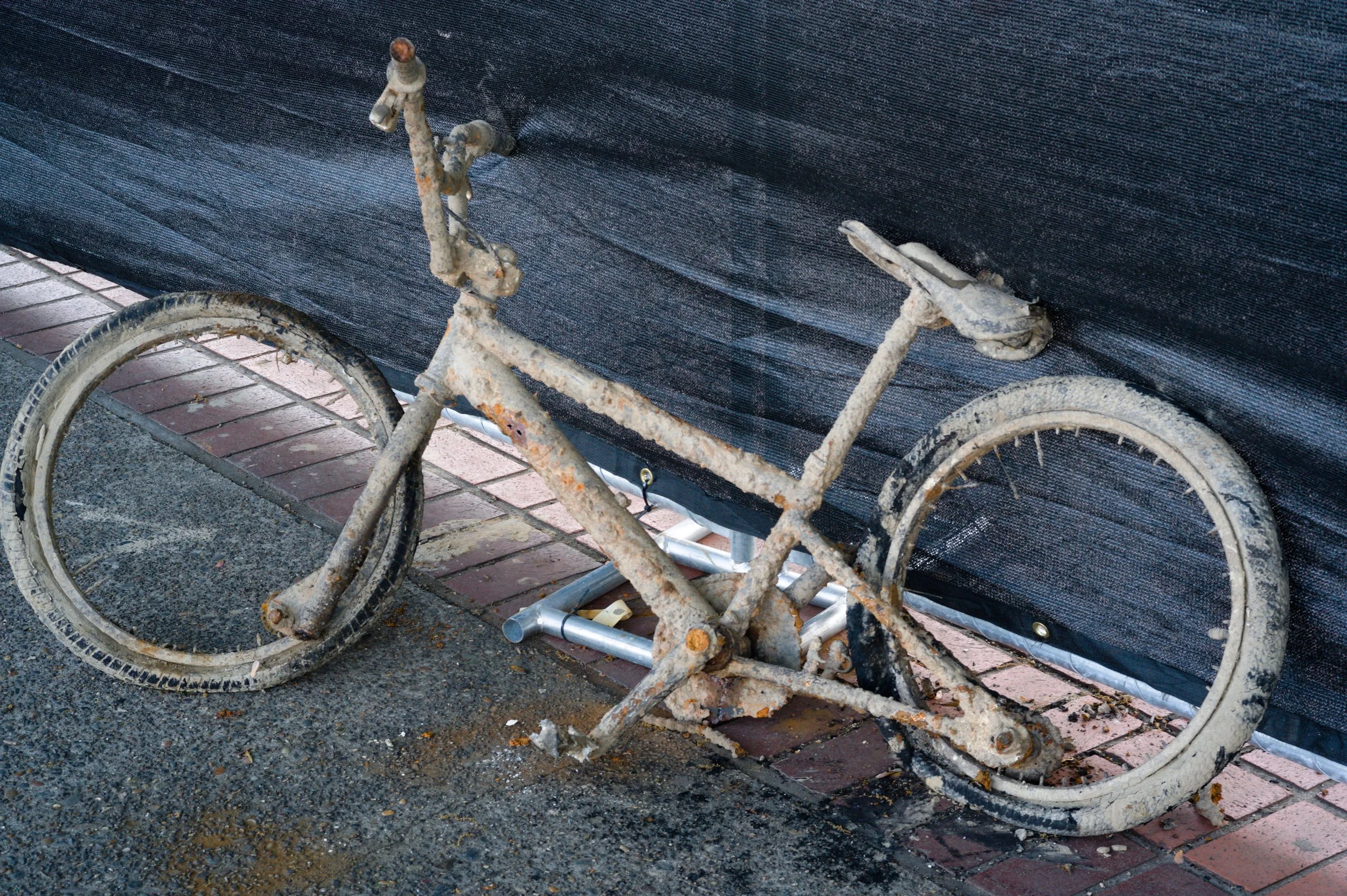Rusty, abandoned bicycle with flat tires and broken parts, leaning against a black fabric backdrop on a sidewalk.