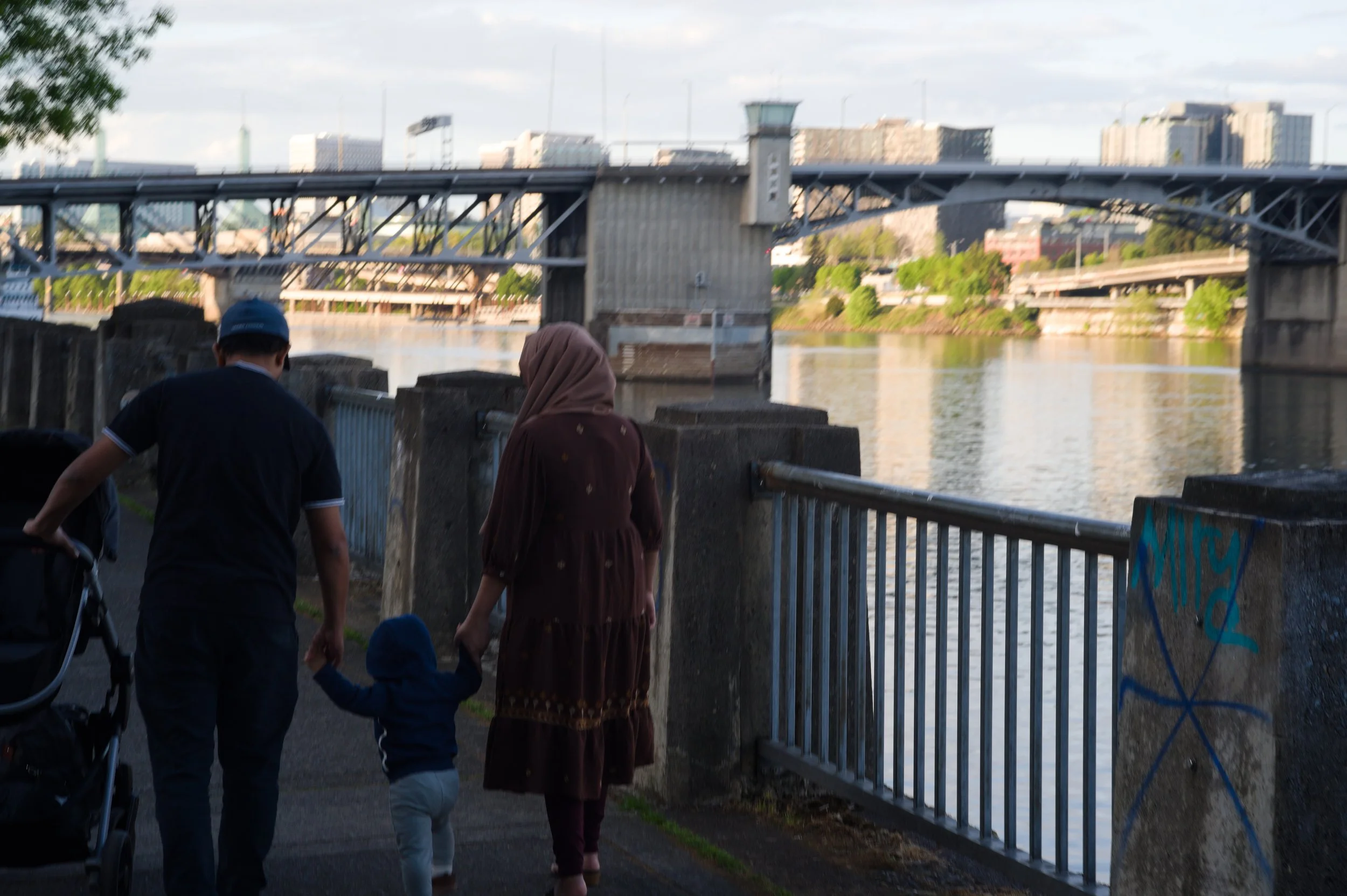 People walking along a riverside walkway near a bridge with an urban skyline in the background.
