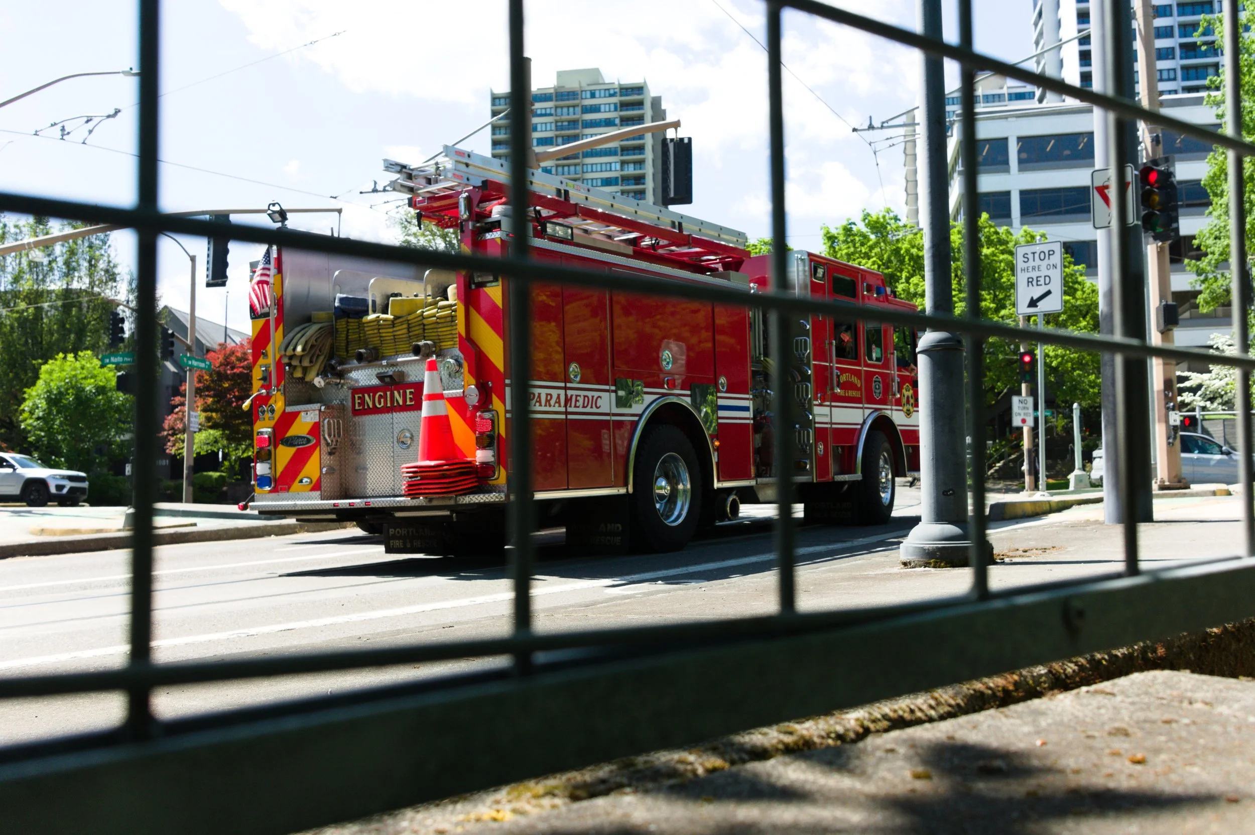 A red fire truck is on the street, seen through a fence with city buildings, trees, and traffic lights in the background.