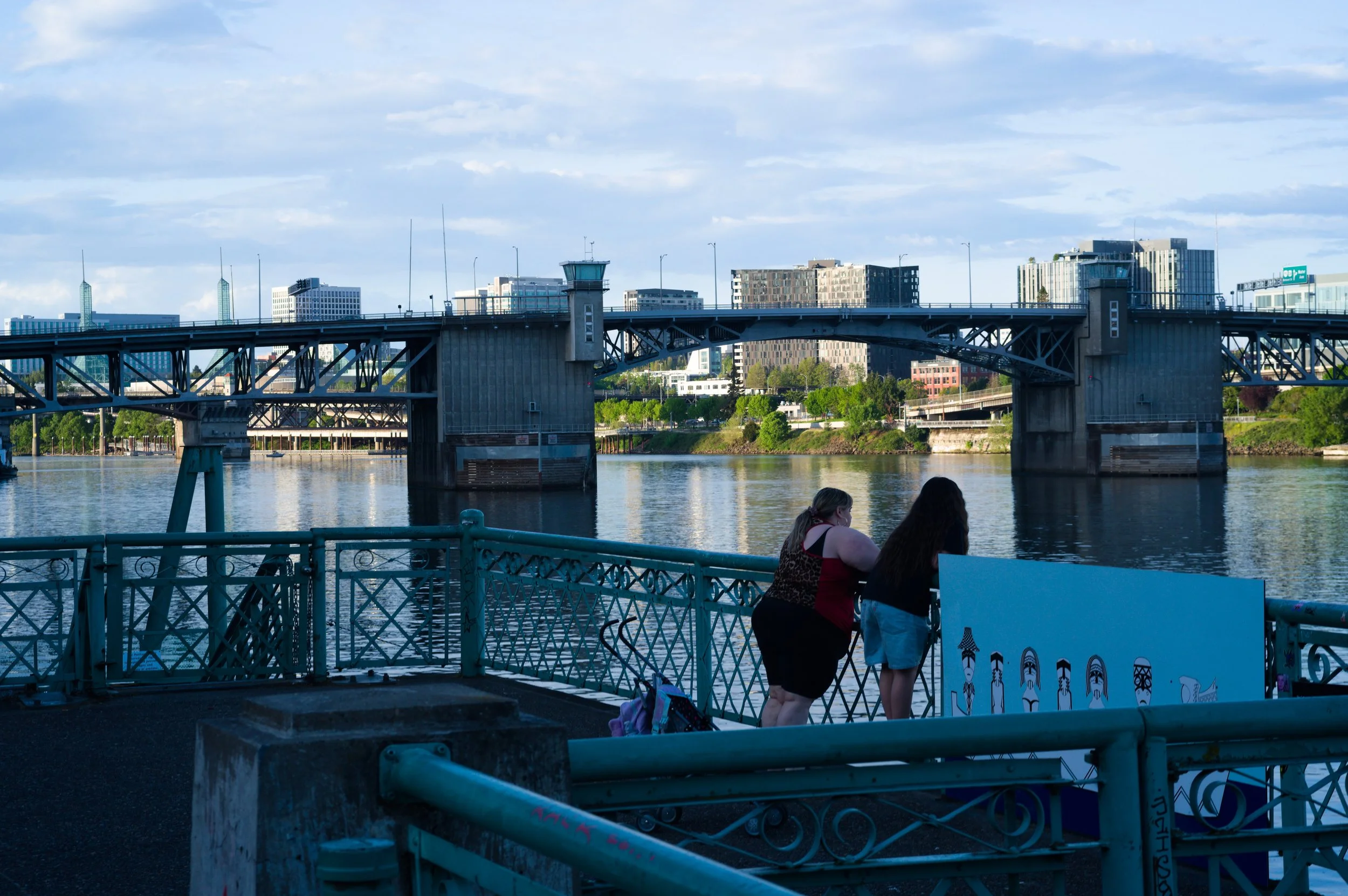 People walking along a riverwalk with a bridge and cityscape in the background.