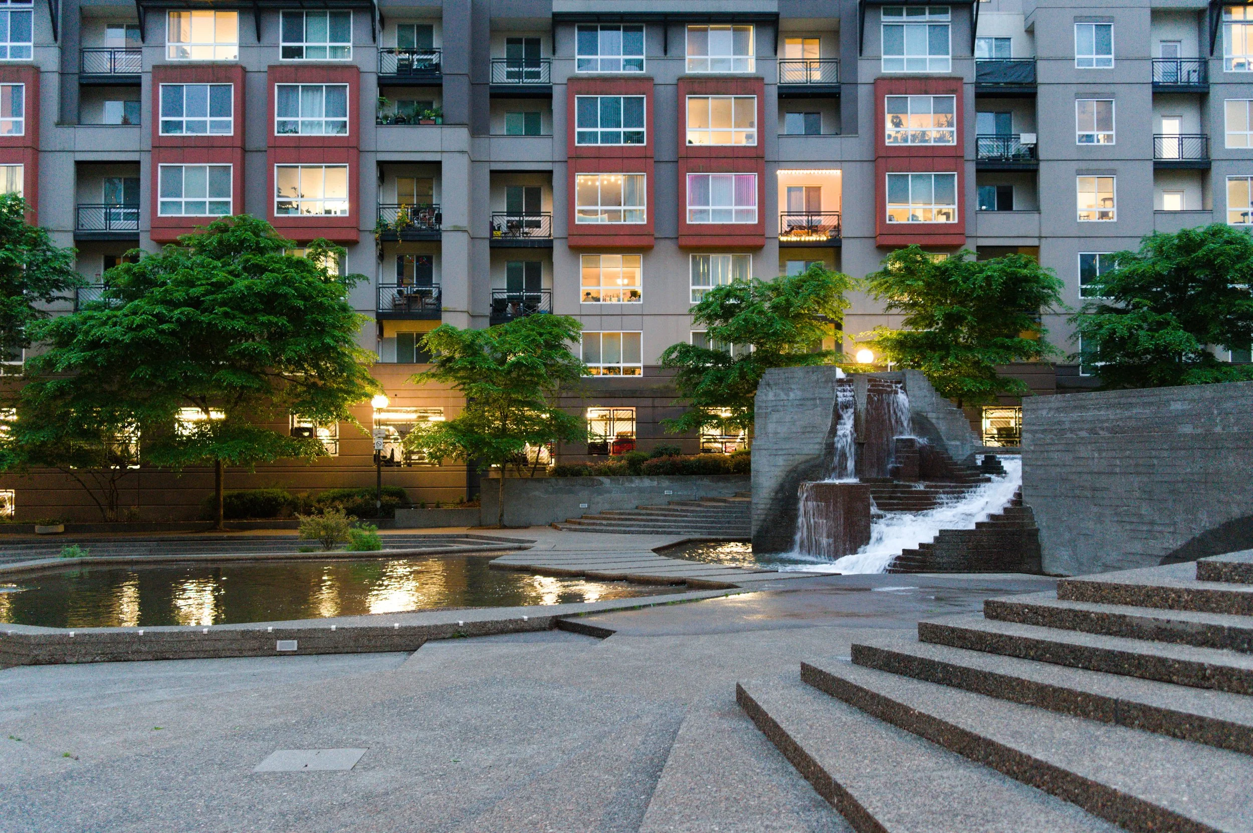 An apartment building with multiple lit windows, a landscaped area with trees, a water feature, and steps leading up to the building at dusk.