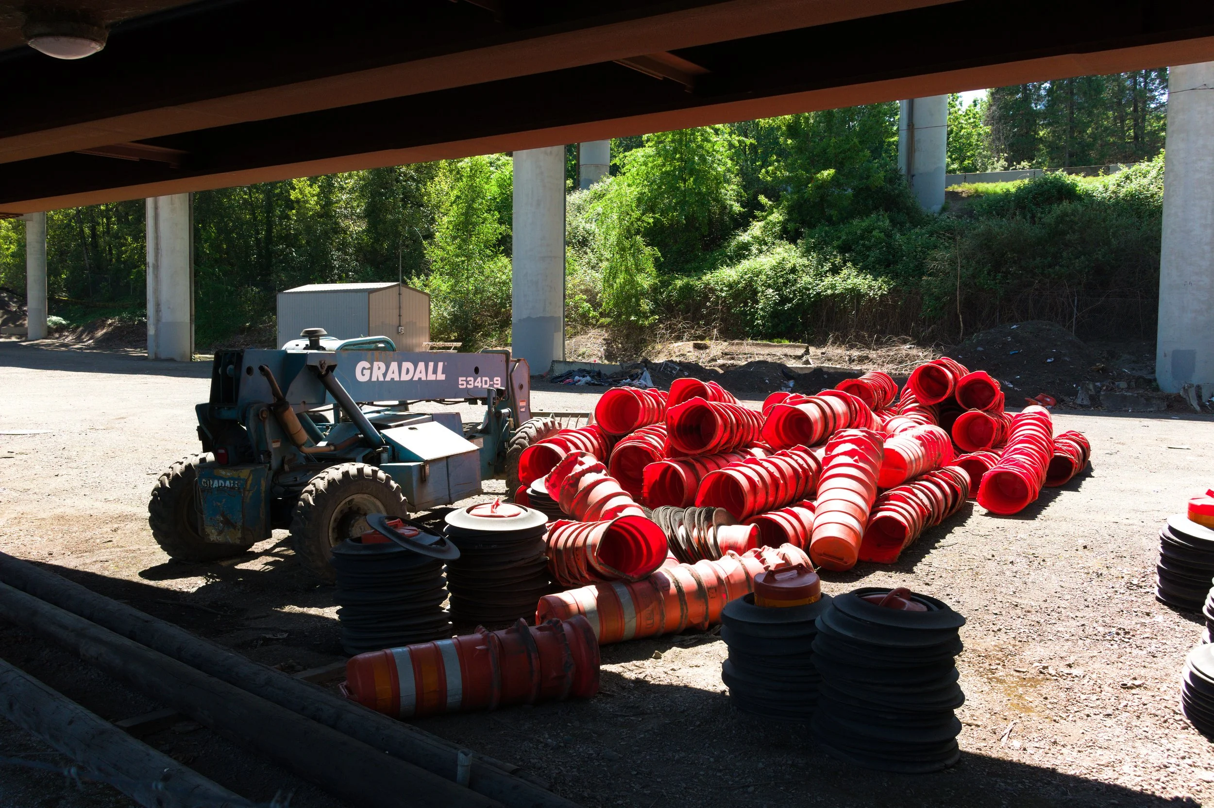 Construction site with orange traffic cones scattered on the ground and a small blue bobcat in the foreground. Under the bridge, the area is shaded while bright green trees and bushes are visible in the background.