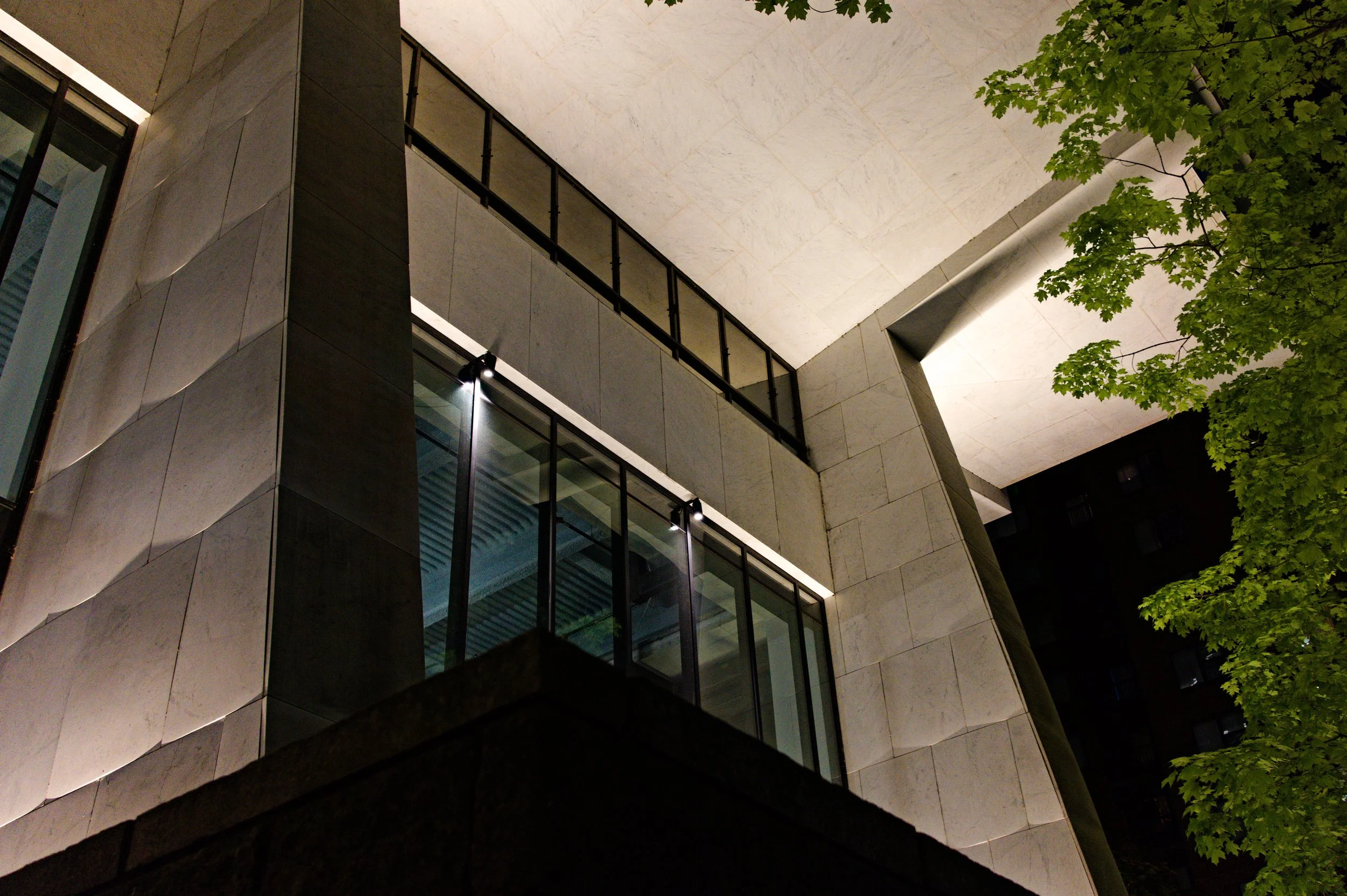 Night view of a modern building with glass windows, metal railings, and concrete walls, partially obscured by green trees.