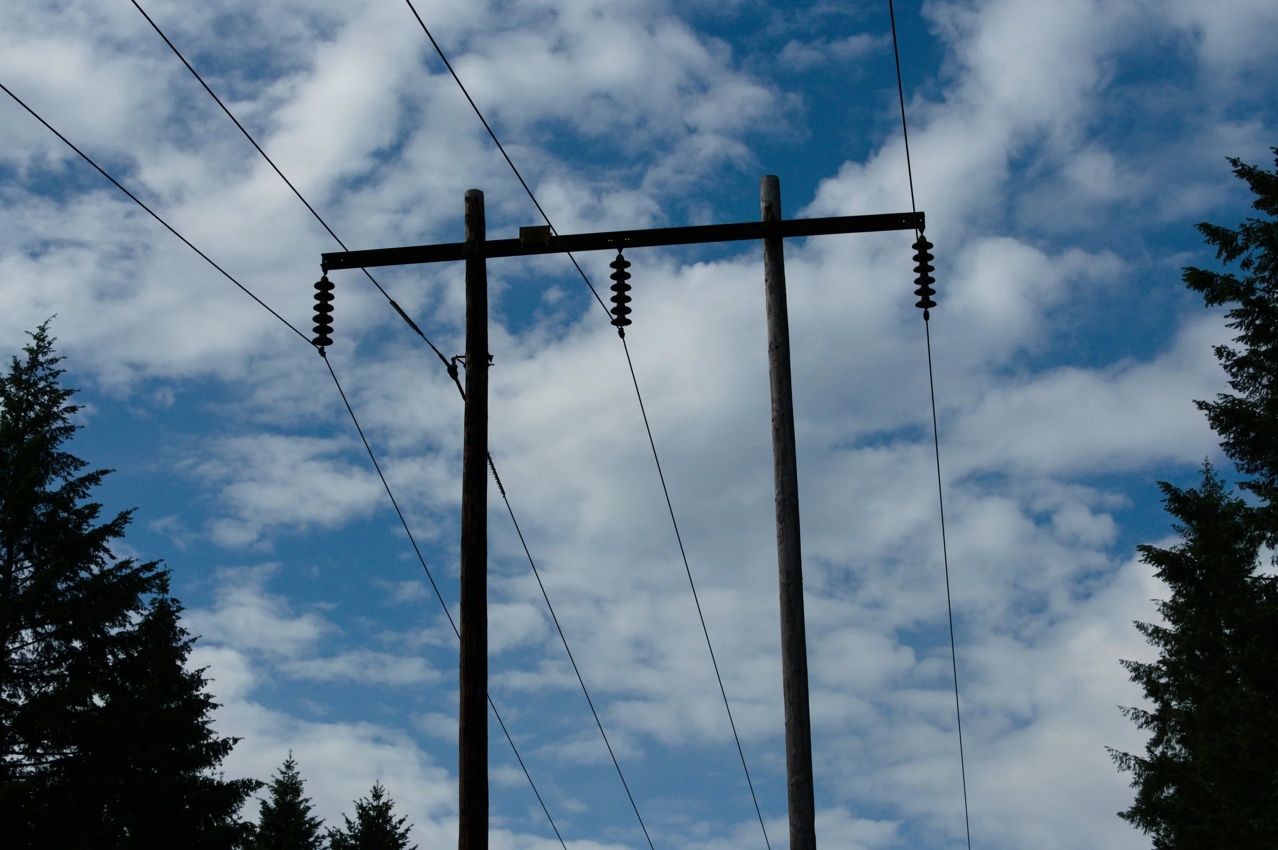 Photograph of power lines and utility poles against a partly cloudy sky with some tree branches visible.