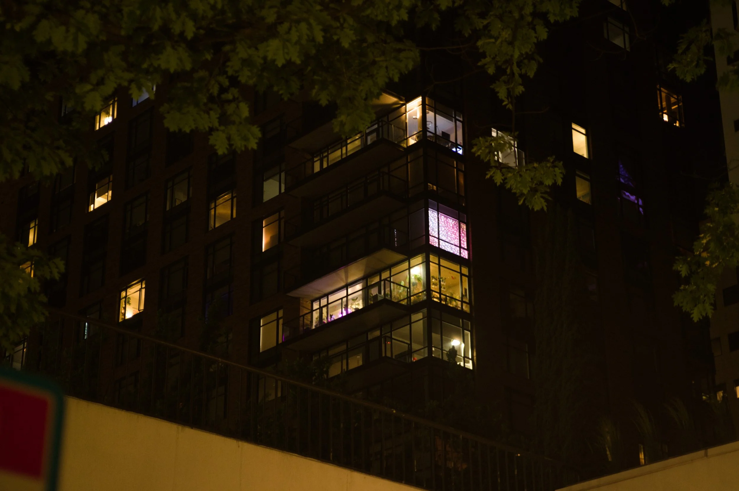 Night view of a multi-story apartment building with illuminated windows, partially obscured by tree branches.