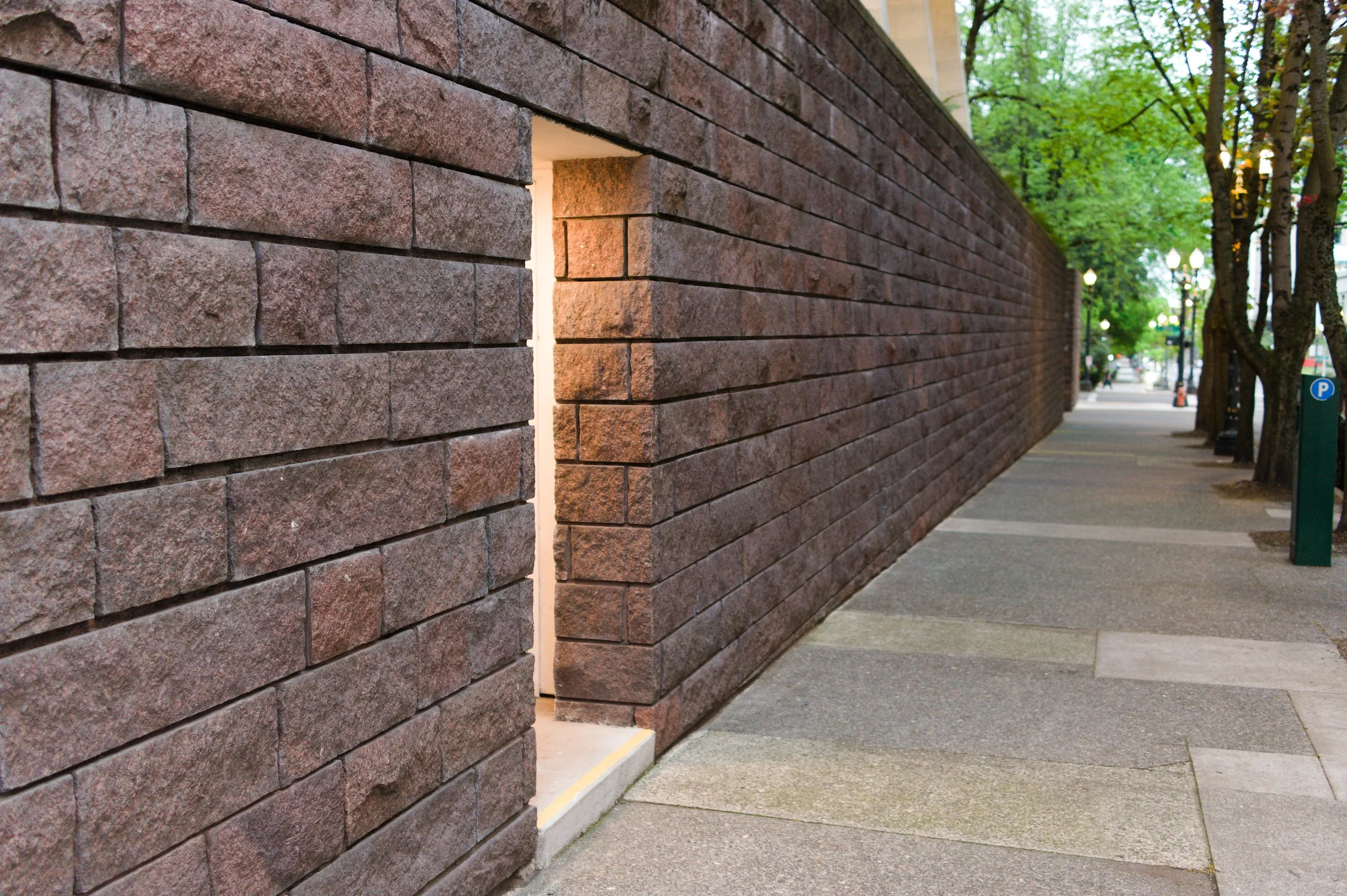 A brick wall with a small opening or window in the sidewalk, with trees and street lamps visible in the background.