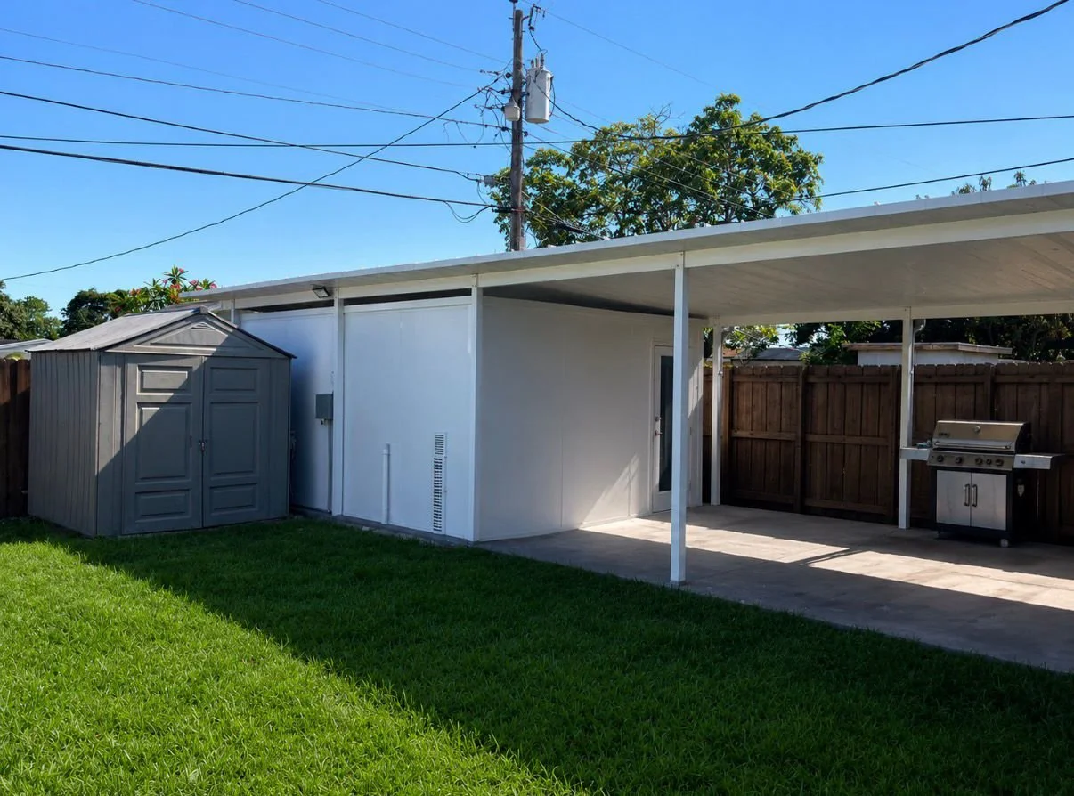 Backyard with a gray shed, a covered patio area with a grill, a white wall, and a wooden fence; green grass and a clear blue sky.