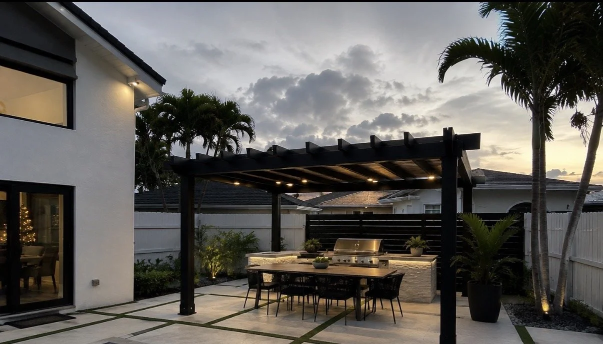 An outdoor backyard kitchen and dining area with a black pergola, a built-in grill, and a table with six chairs, surrounded by a white fence, palm trees, and landscaping at sunset.