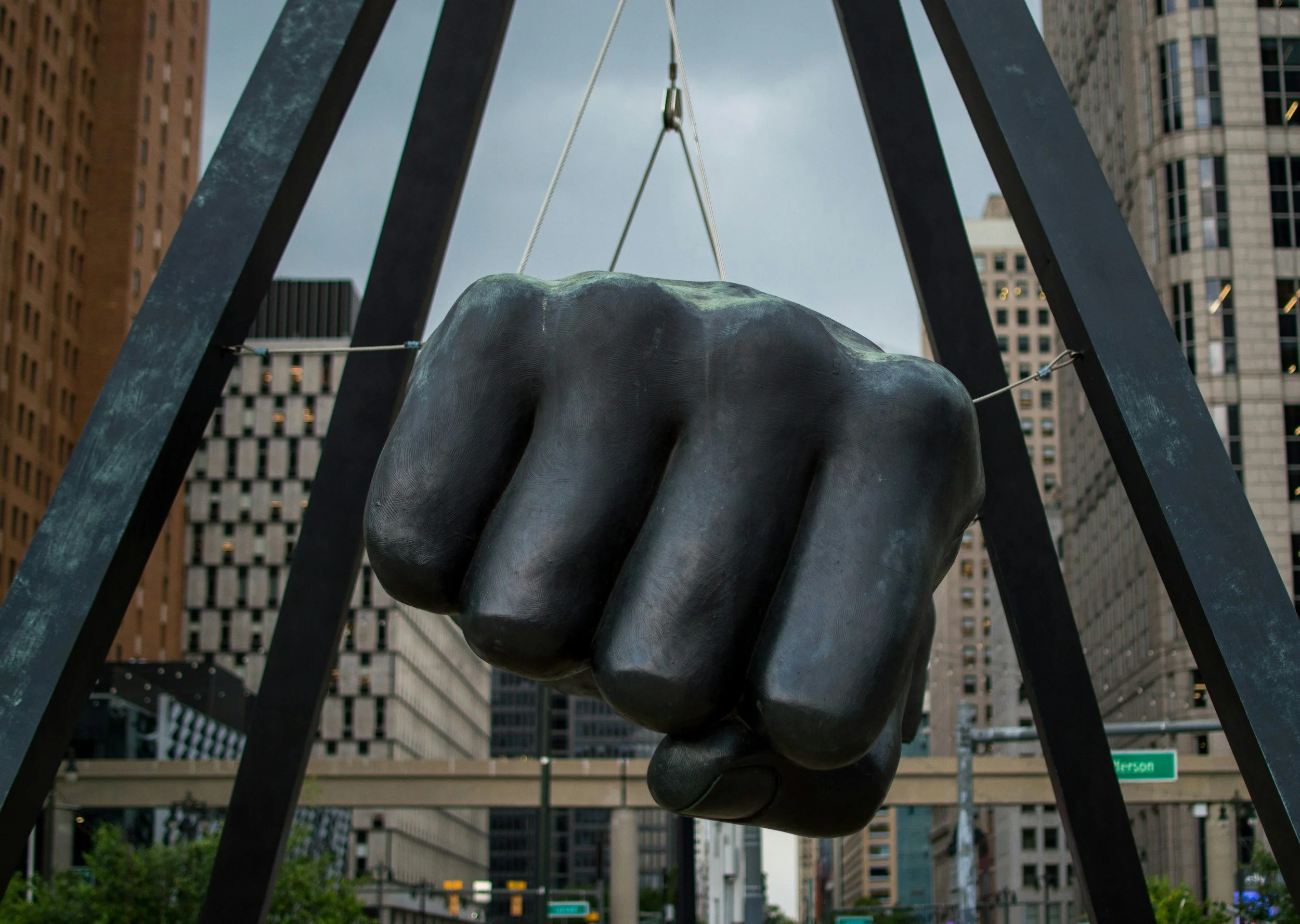 Close-up of a large bronze sculpture of a fist hanging by ropes between black metal beams, with tall city buildings in the background.