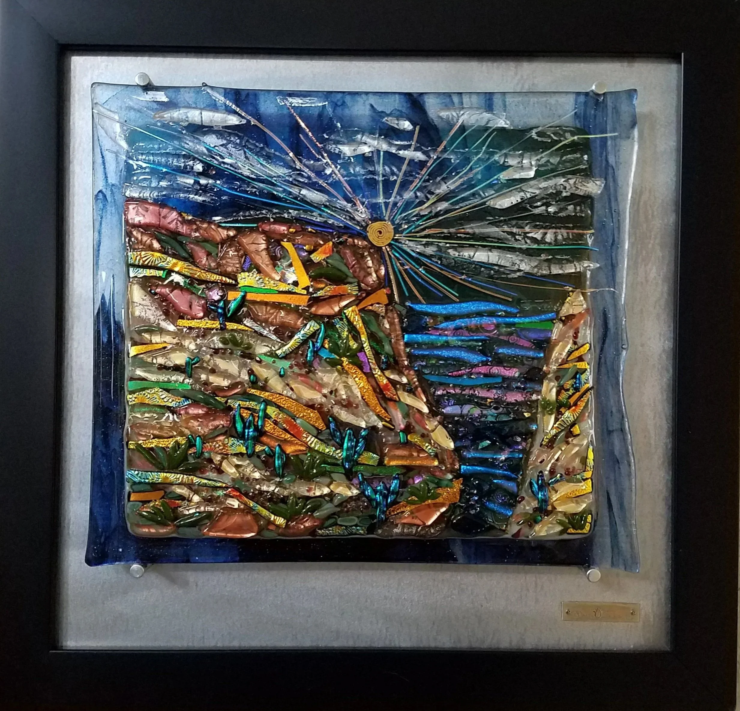 Colorful glass art framed on a wall, depicting a stylized peacock with vibrant blue, green, and gold feathers.