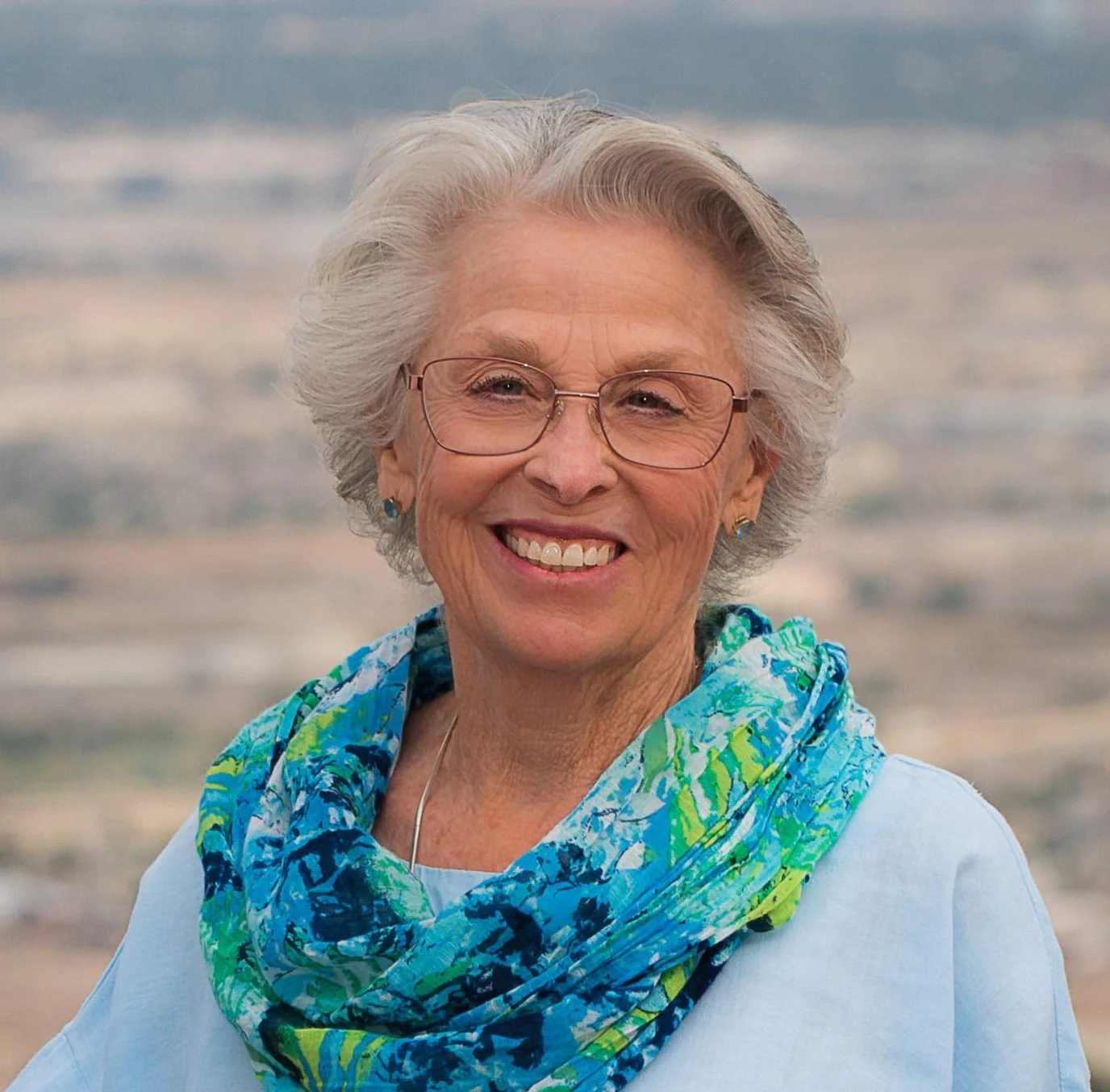 Smiling elderly woman with glasses and short gray hair, wearing a light blue shirt and a colorful blue-green scarf, standing outdoors with a beach and ocean in the background.