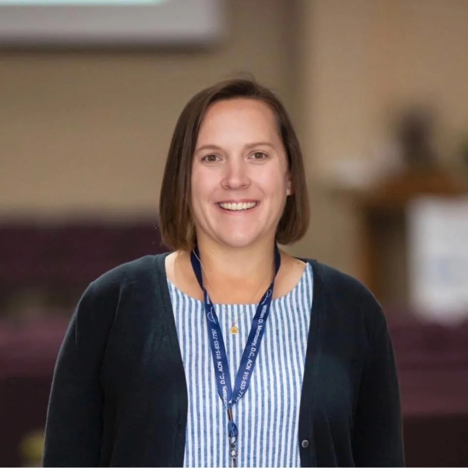 Smiling woman with shoulder-length brown hair wearing a blue lanyard, striped shirt, and black cardigan.