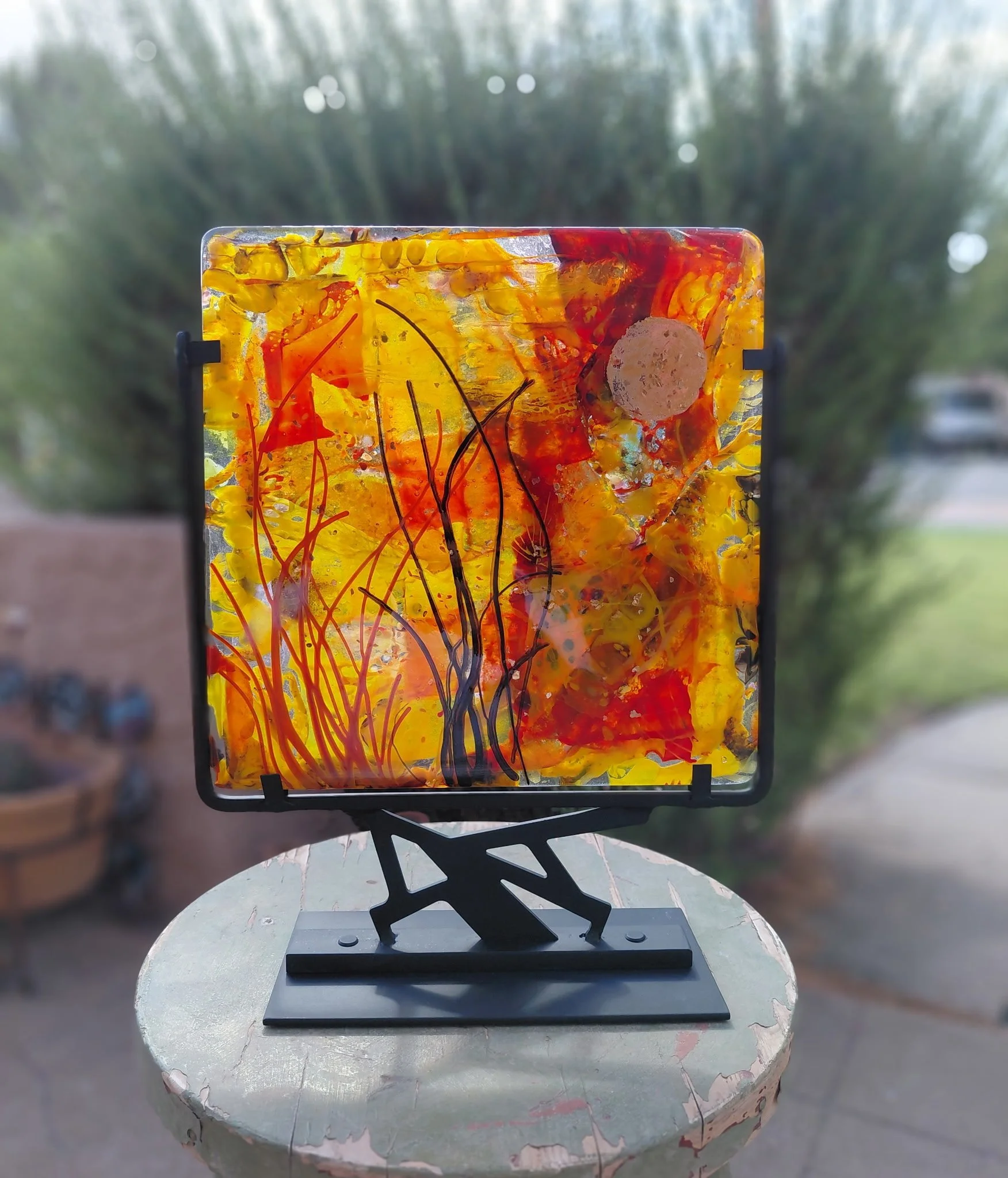 Colorful abstract art piece with yellow, orange, red, and black streaks on glass, displayed on a black stand outdoors on a white round table.
