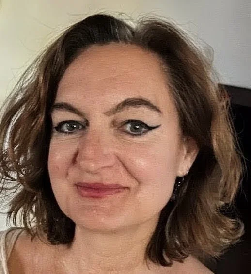 Close-up of a woman with wavy brown hair, blue eyes, winged eyeliner, and pink lipstick, smiling at the camera.