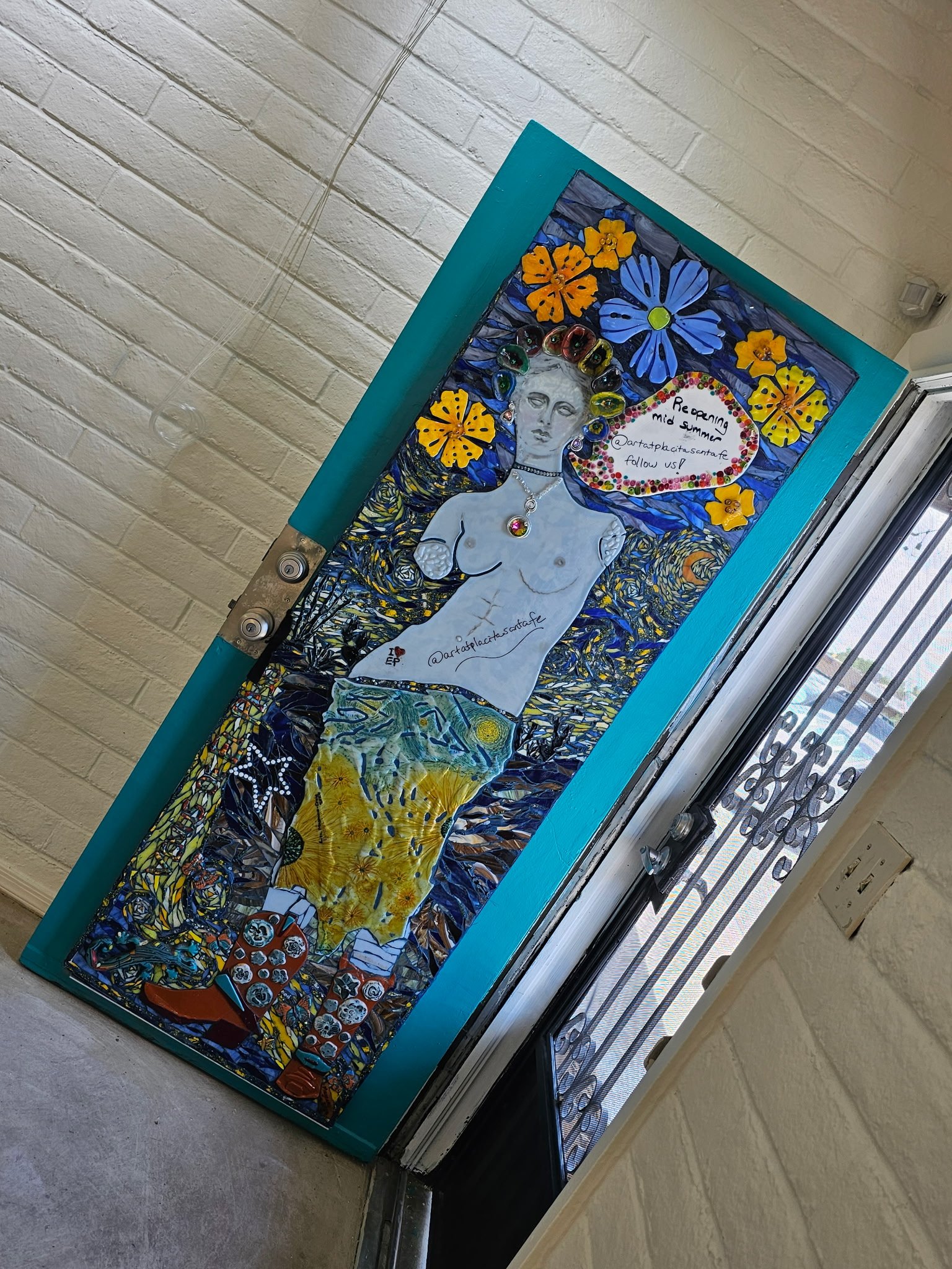 Colorful door decorated with a mosaic of flowers, a portrait of a woman with jewelry, and a sign that says 'Reopening mid summer @artatpacitasantafe follow us!'. The mosaic features vibrant yellow and blue flowers and a painting of a woman with a necklace, with a backdrop of swirling blue and yellow patterns.