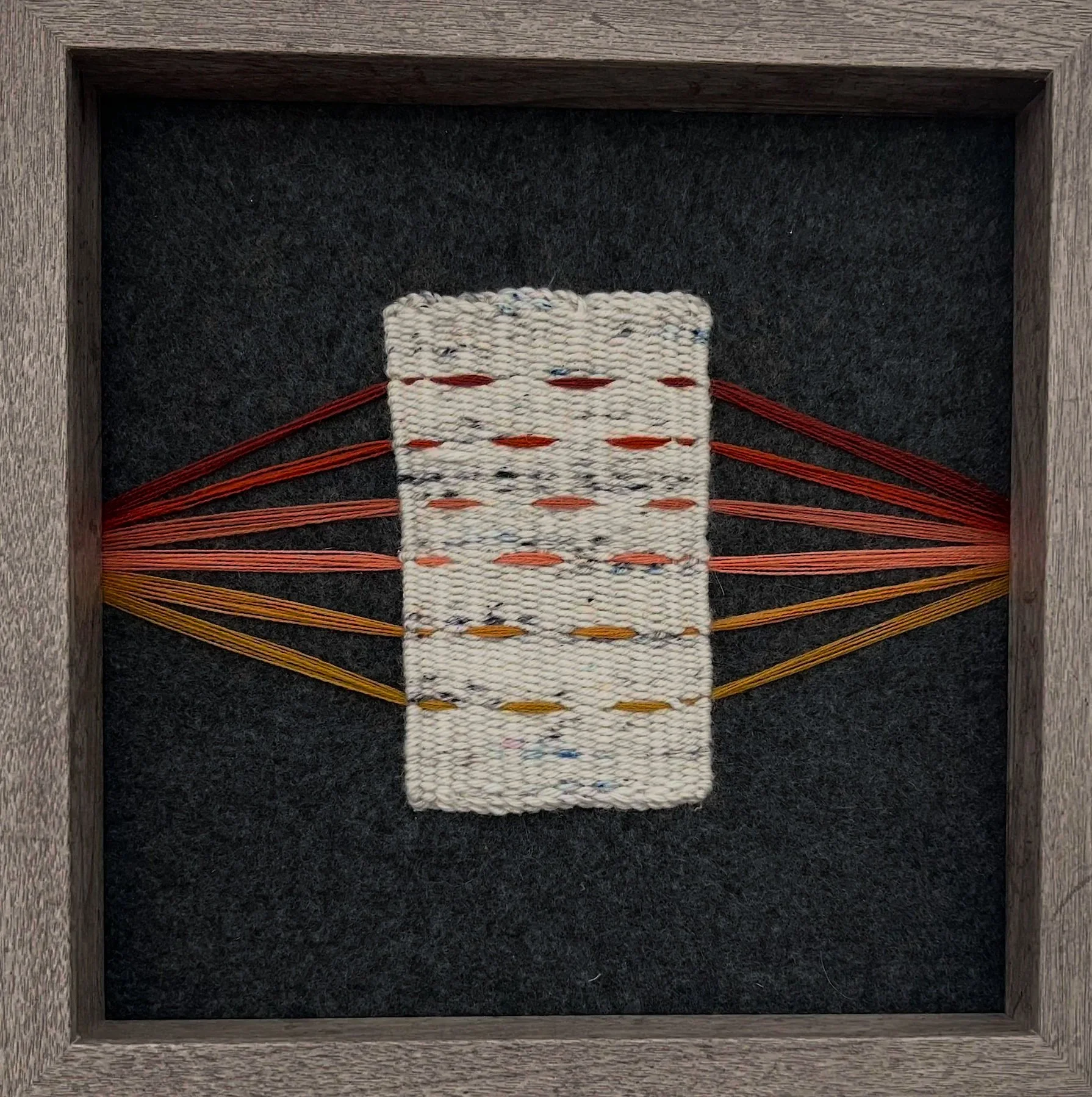 A handmade woven rug with orange, yellow, and black threads, placed inside a wooden shadow box frame on black fabric background.