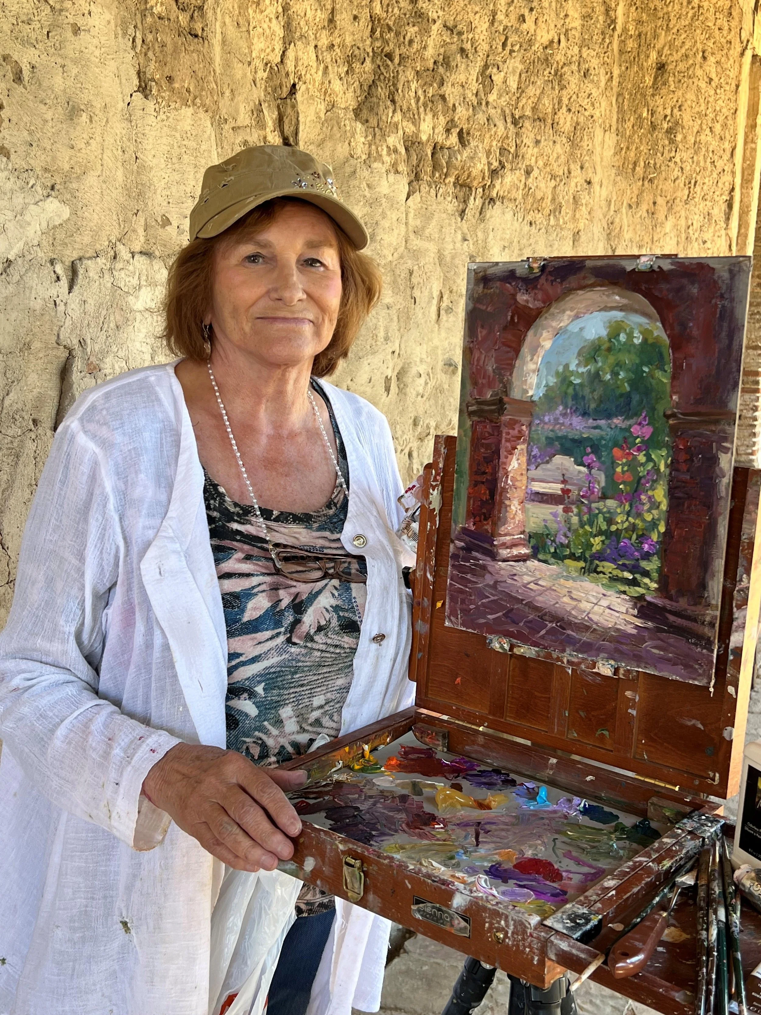 A woman with short red hair, wearing a beige hat and a white jacket, is standing next to an easel with a painting of an arched stone gateway on it. The scene appears to be outdoors with a yellowish textured wall in the background.