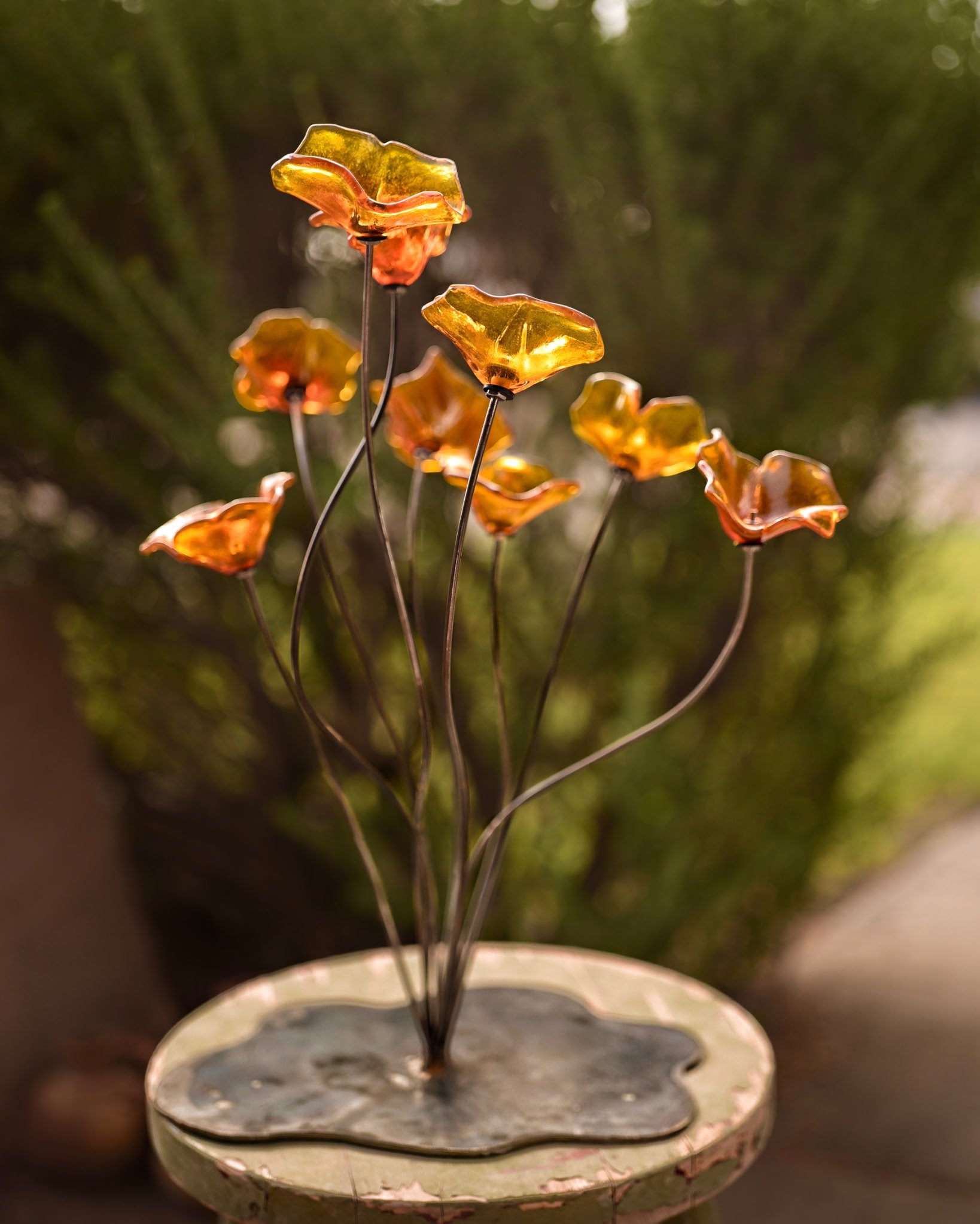 A decorative silver tray with a floral-shaped design, holding a display of lighted amber-colored glass flowers on thin metal stems, placed against a blurred green natural background.