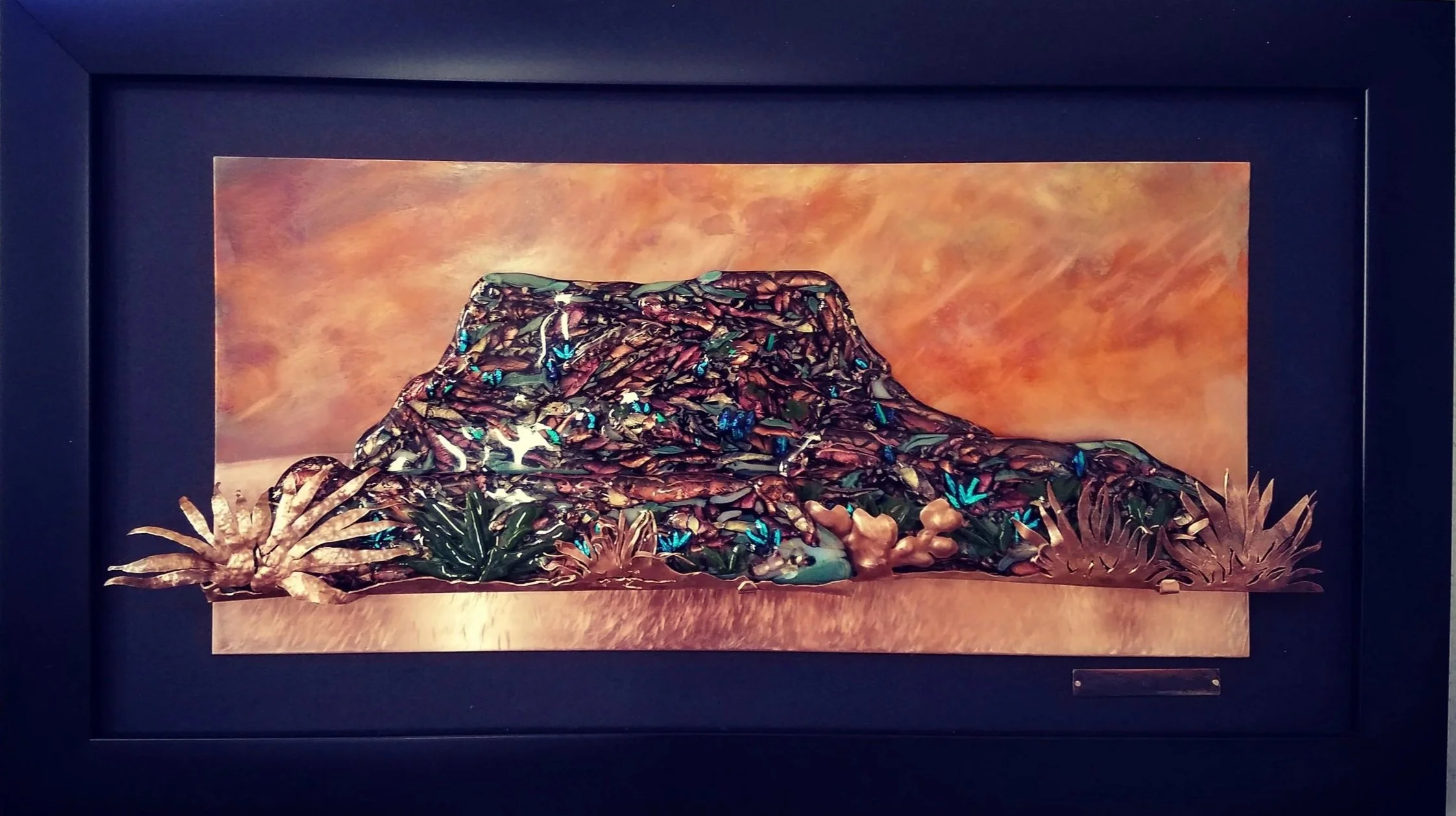 a framed painting of a colorful desert landscape with layered rocks, desert plants, and a vibrant sky in shades of orange and pink.