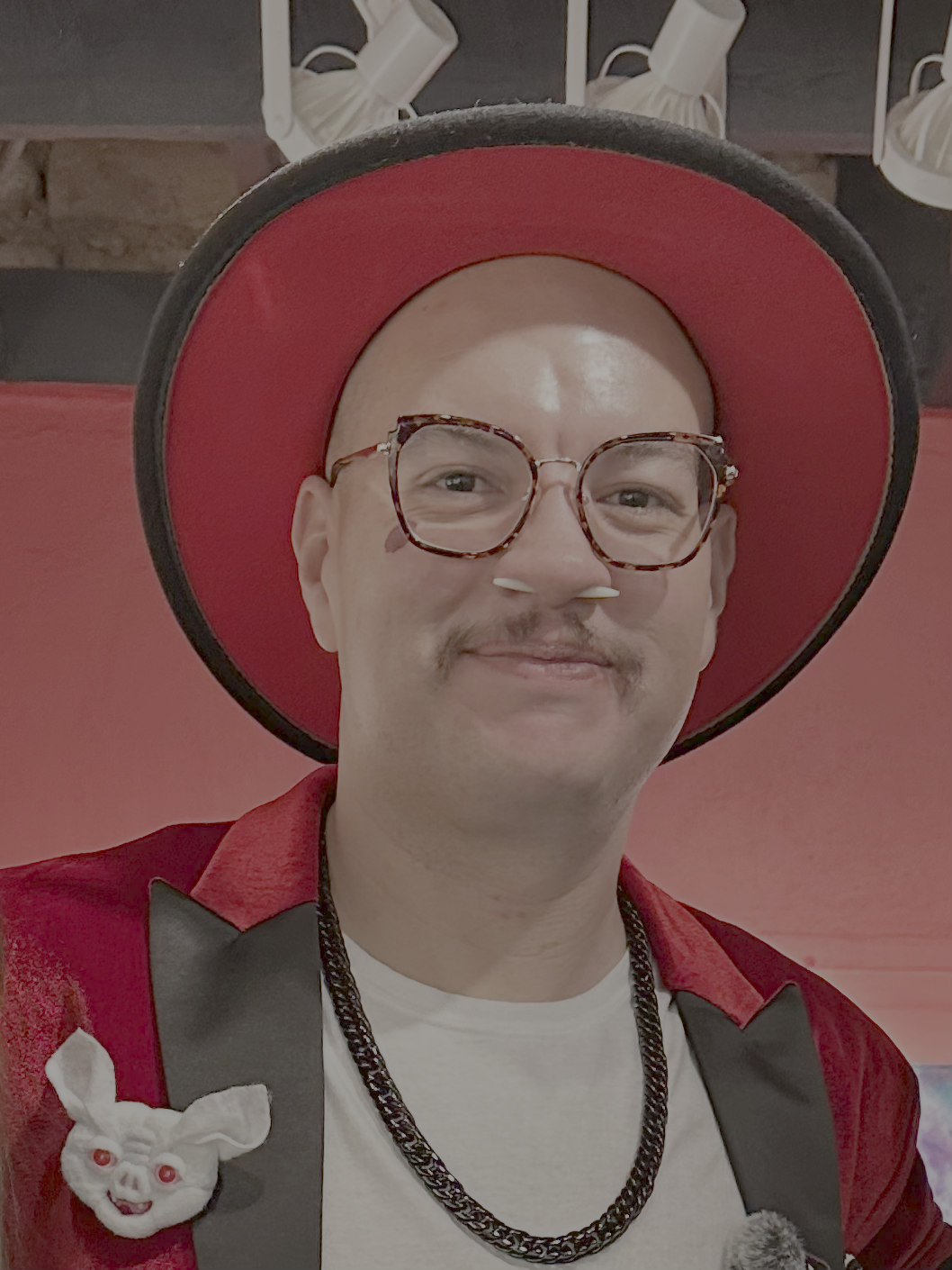 A person wearing glasses, a red hat, and a red jacket with a white shirt. They are smiling and have a plush toy pinned to their jacket.