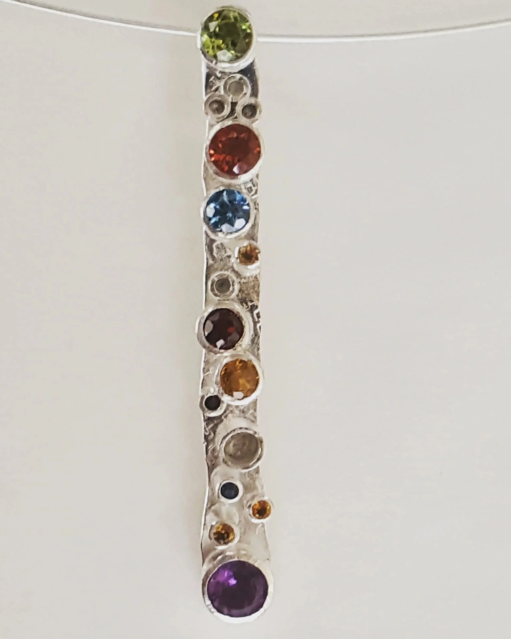 A silver jewelry piece featuring a vertical row of round, multicolored gemstones, including green, brown, blue, purple, and amber, set in individual circular settings.
