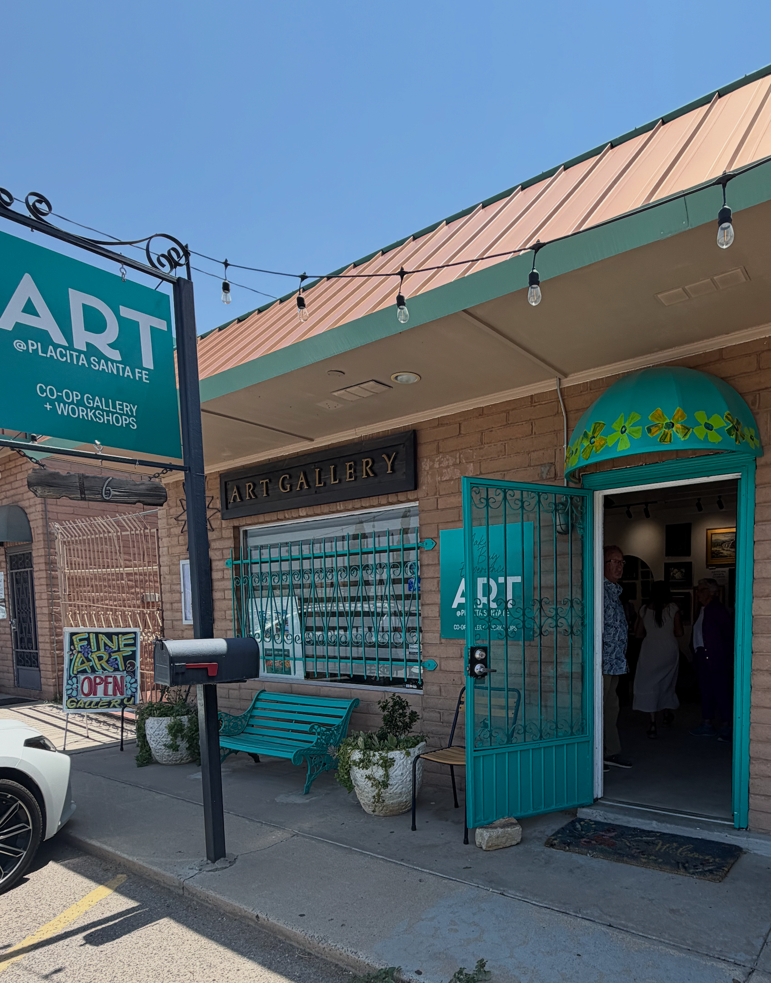 The exterior of an art gallery with teal-colored door and window frame, decorated with string lights, a teal bench, and potted plants. People are entering the gallery.