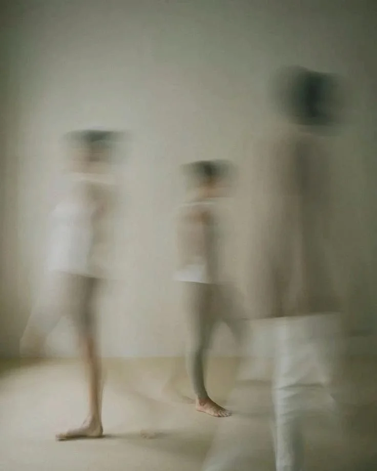 Three people walking in a room, with two women in white dresses and one person dressed in white pants and a white top, in front of a white wall and some furniture.