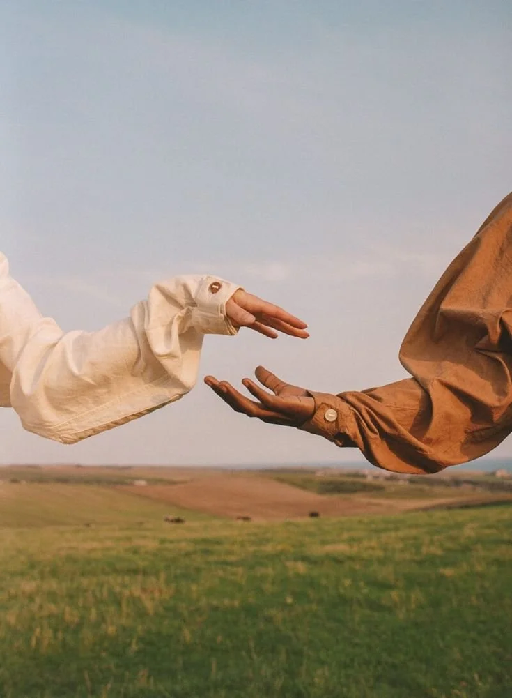 Two hands reaching out towards each other in an open field under a cloudy sky.