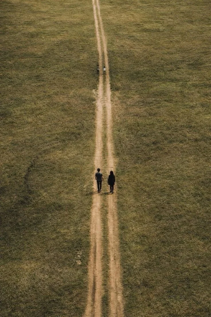 An aerial view of a grassy field with a dirt path running through the middle. A couple is walking together on the path near the foreground, and a person is walking further ahead in the distance.